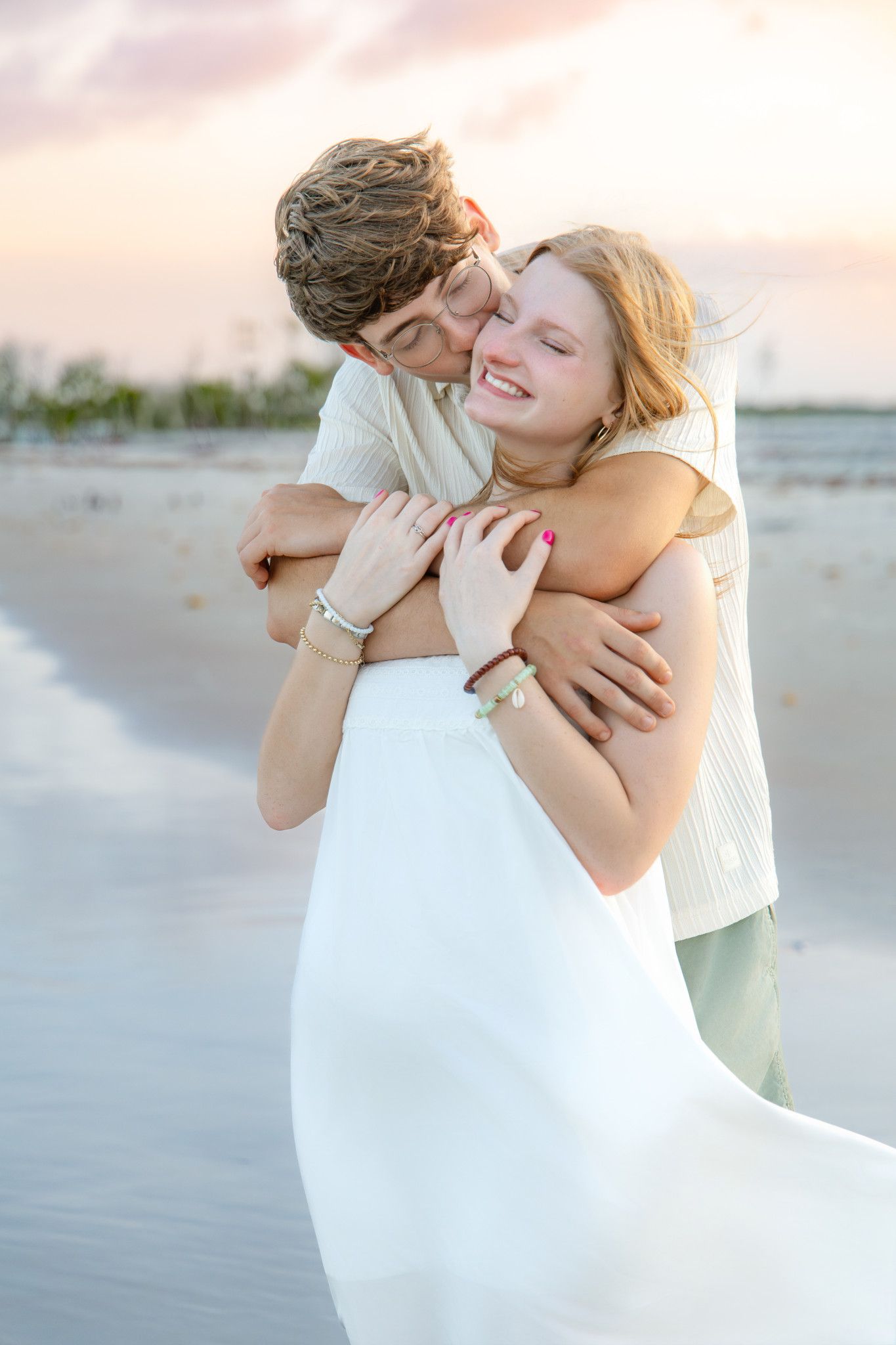 Couple embracing on beach at sunset, woman in white dress smiling joyfully as man hugs her from behind.