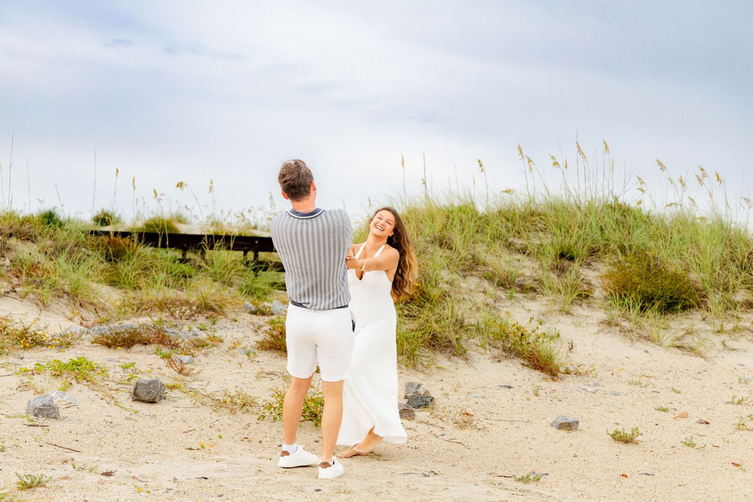 Couple dancing playfully on sandy beach dunes with sea oats in background.