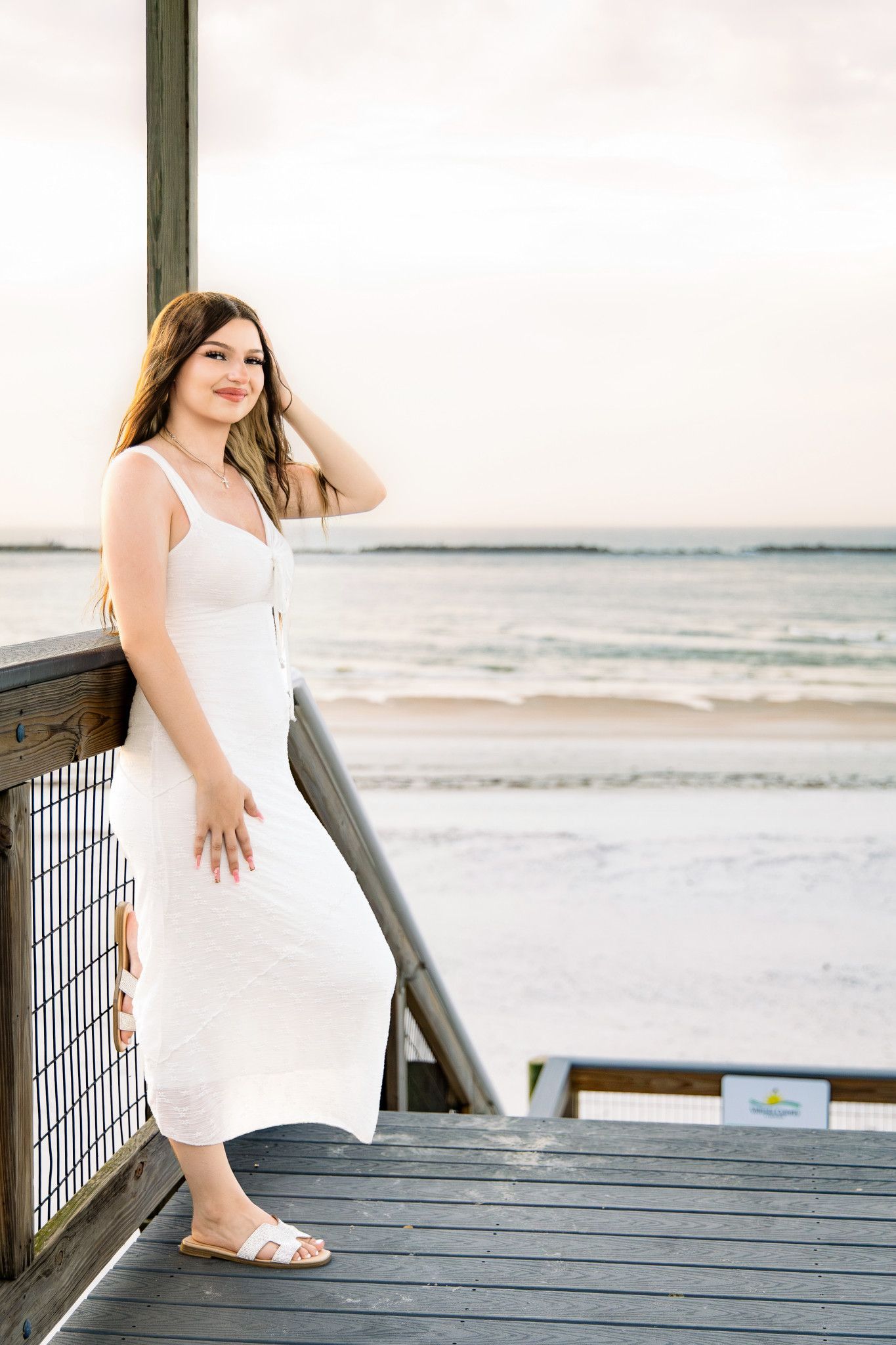 Young woman in white maxi dress posing on a wooden beach boardwalk overlooking the ocean at sunset.