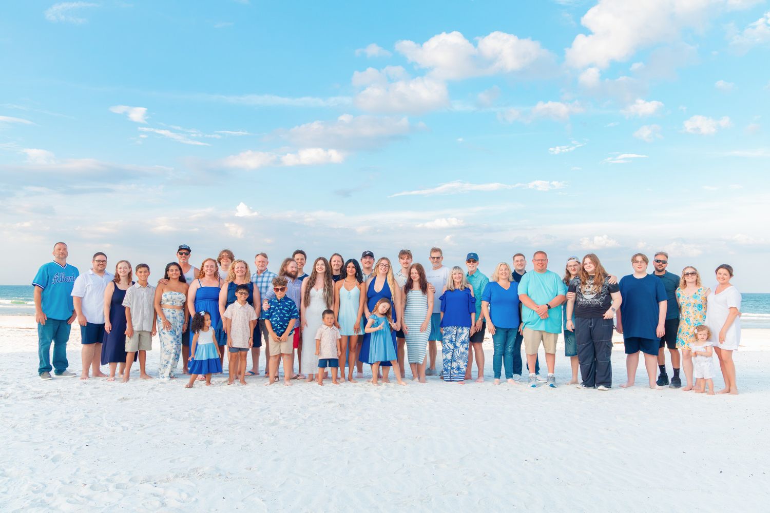 Large family group posing together on a white sandy beach with blue skies and calm ocean waves in the background.