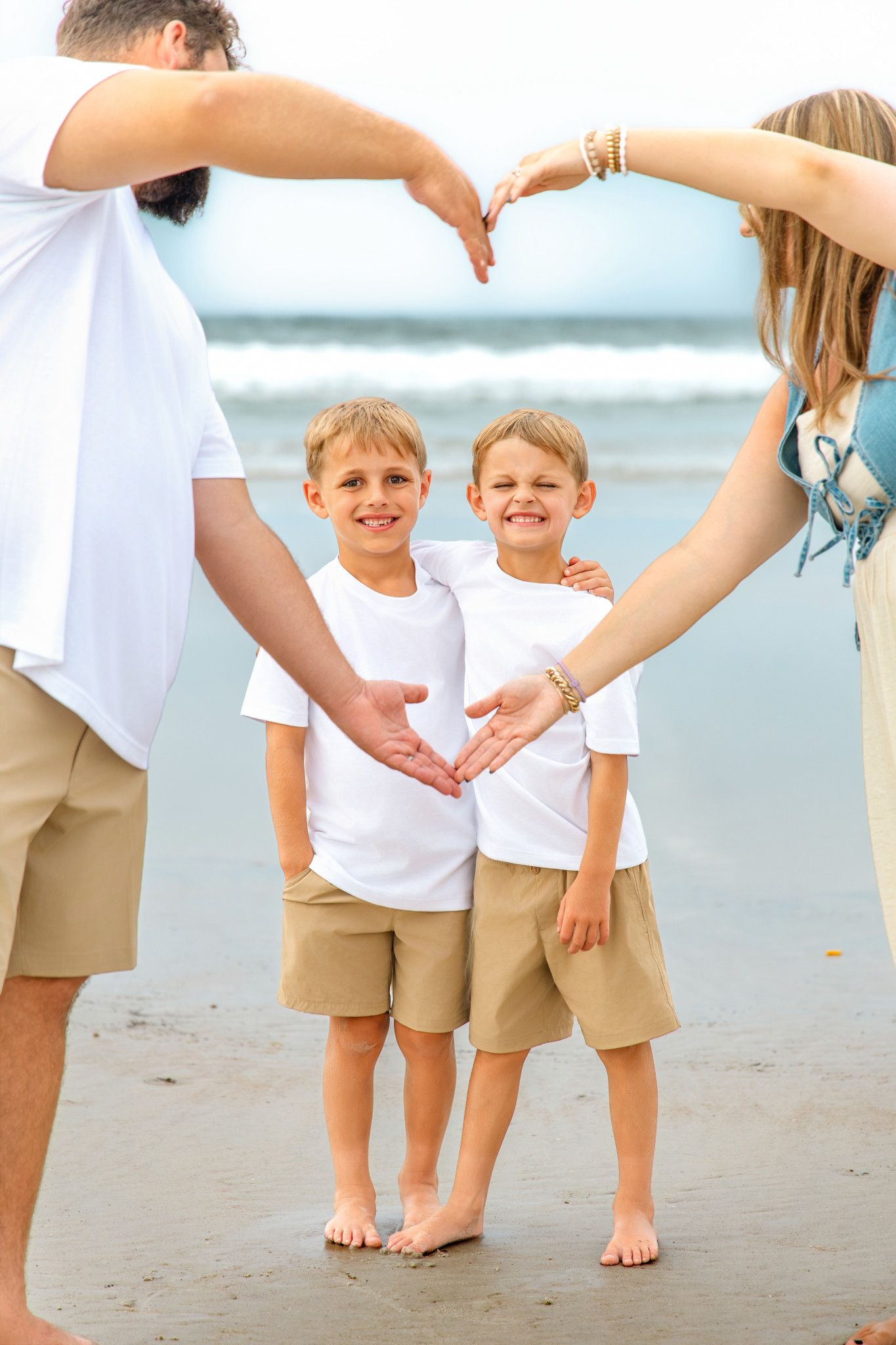 Two smiling twin boys in matching white shirts and khaki shorts on a beach, framed by parents forming a heart with their arms.