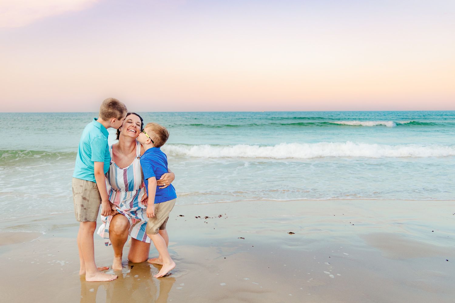 A mother kneels on a sandy beach at sunset while her two young sons lean in to kiss her cheeks near ocean waves.