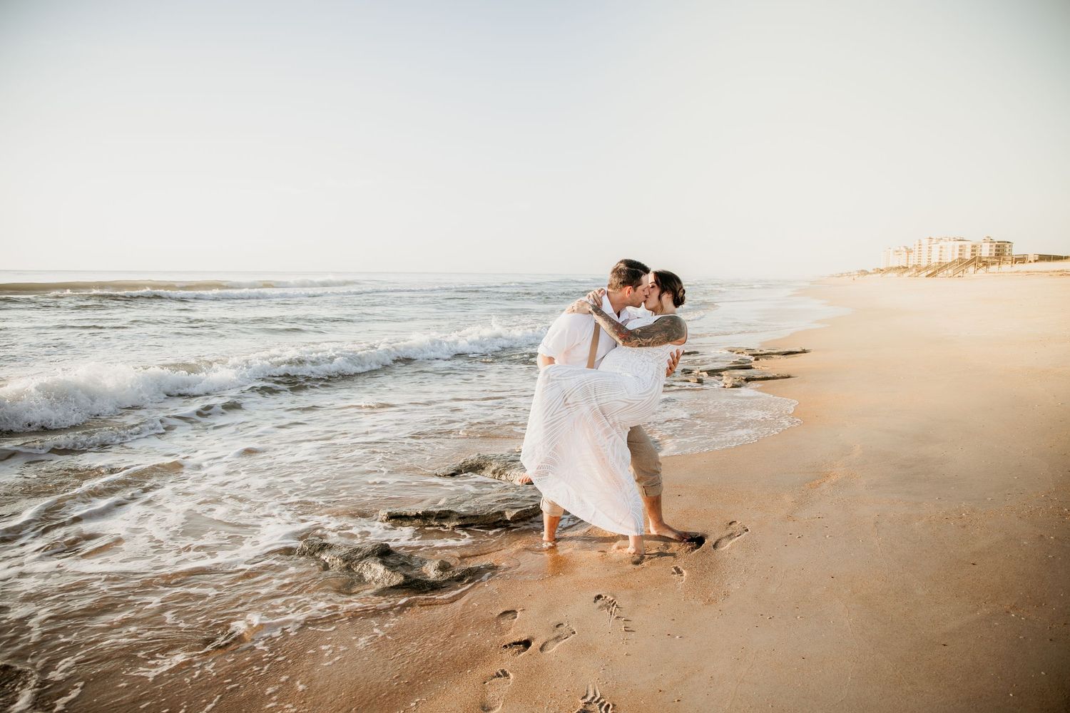 Groom lifts bride in white dress along ocean shoreline during beach wedding.