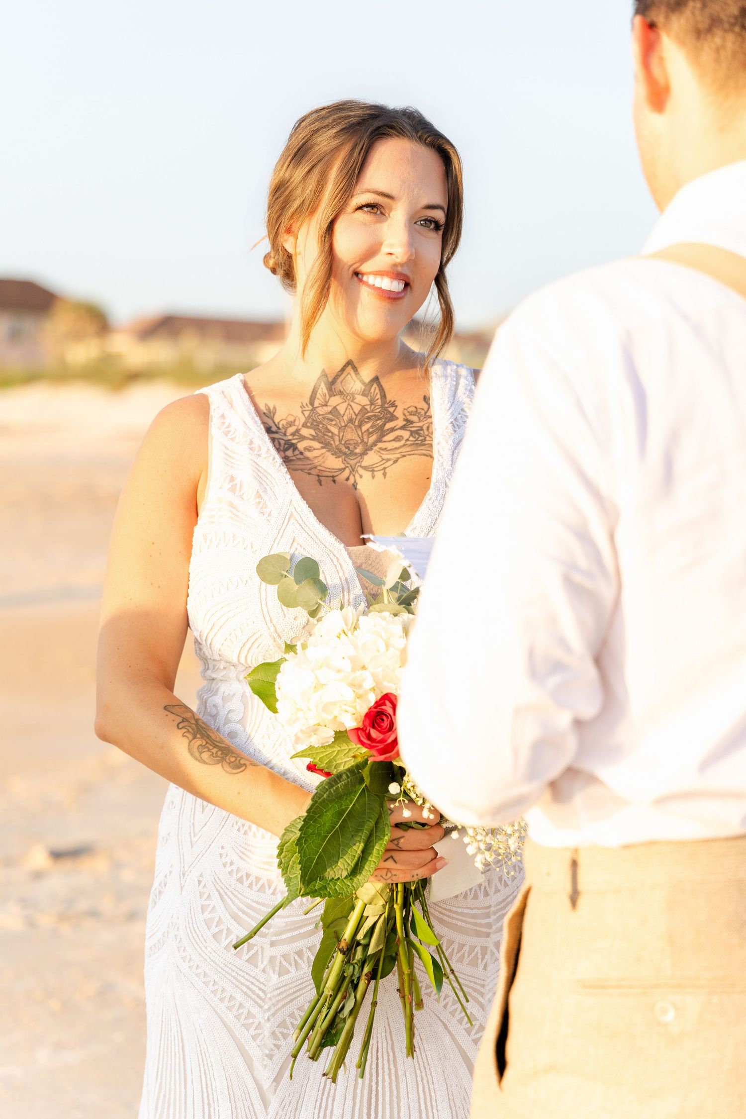 Smiling tattooed bride in lace wedding dress holding colorful bouquet facing groom on sunny beach.