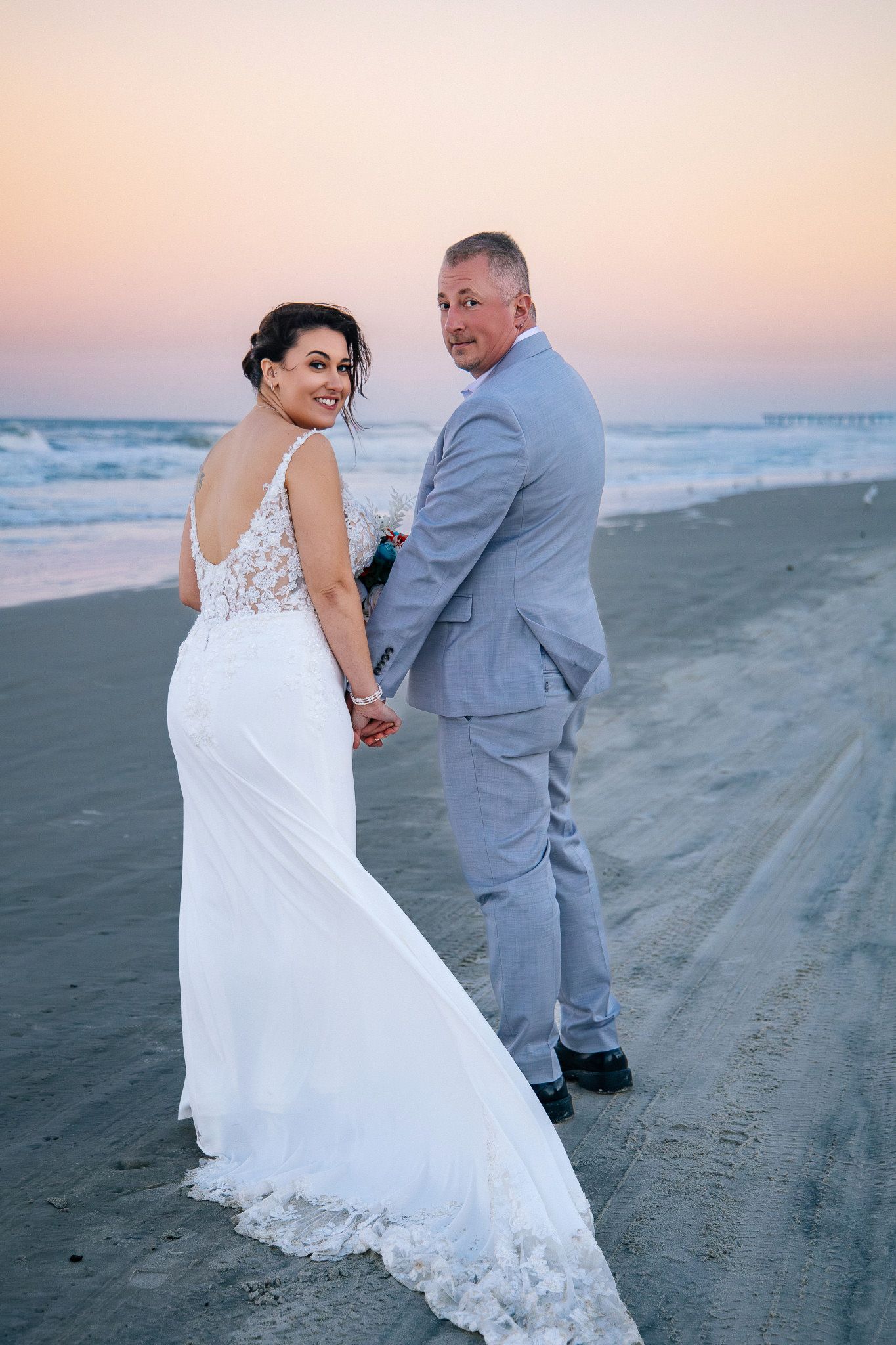 A couple in wedding attire walks along a beach at sunset, with the bride's white lace dress flowing in the breeze.