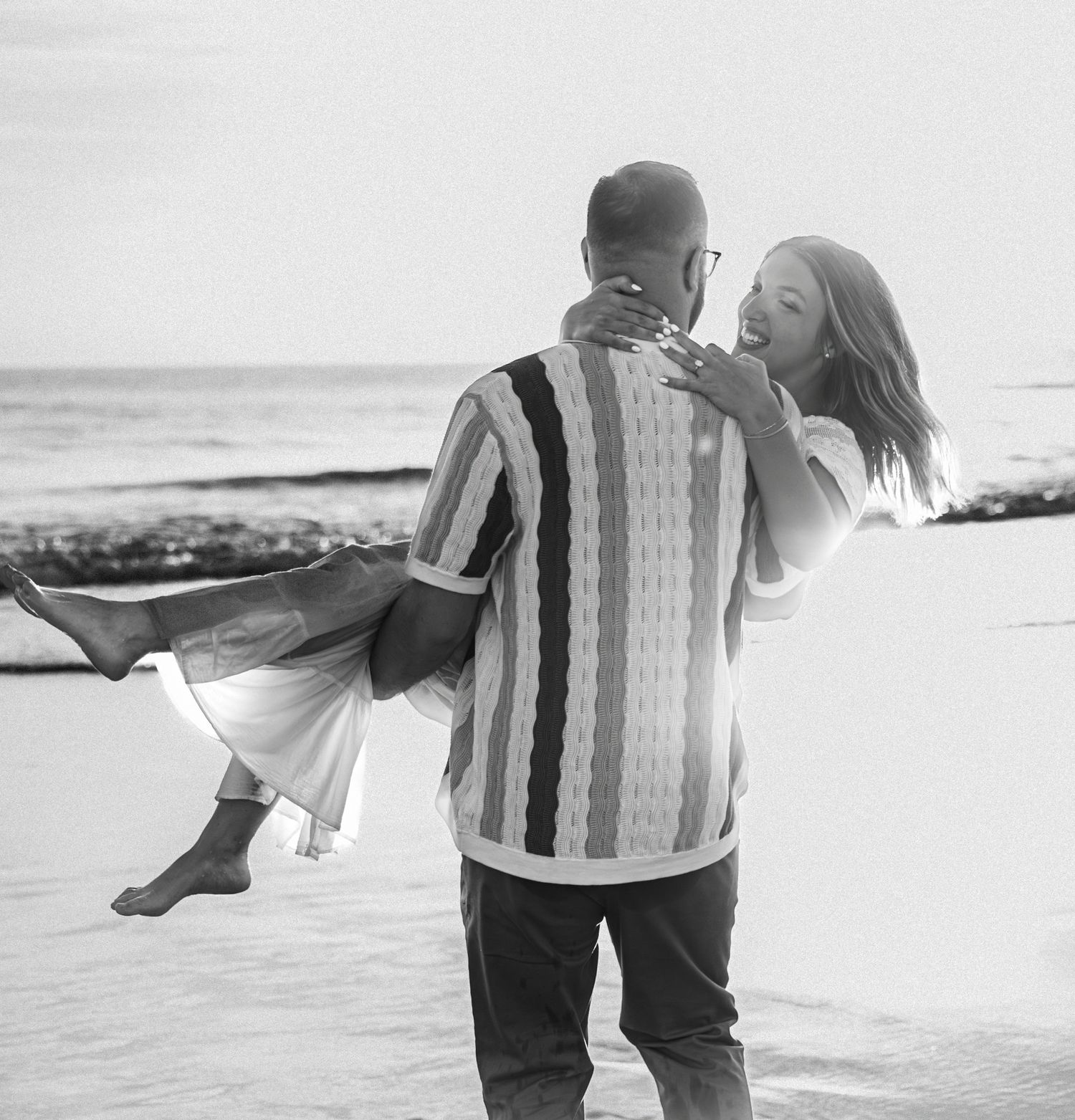 A romantic black and white beach scene shows a couple embracing by the ocean shore.