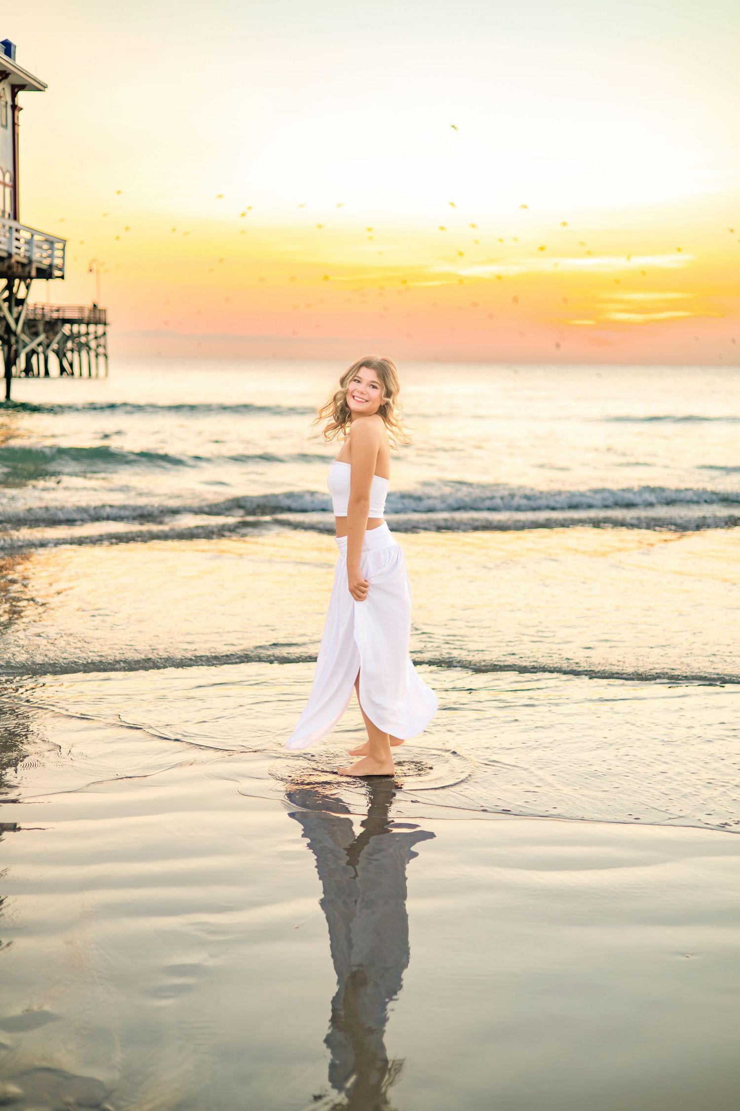 Young woman in white dress walking on beach at sunset with ocean waves and pier in background.