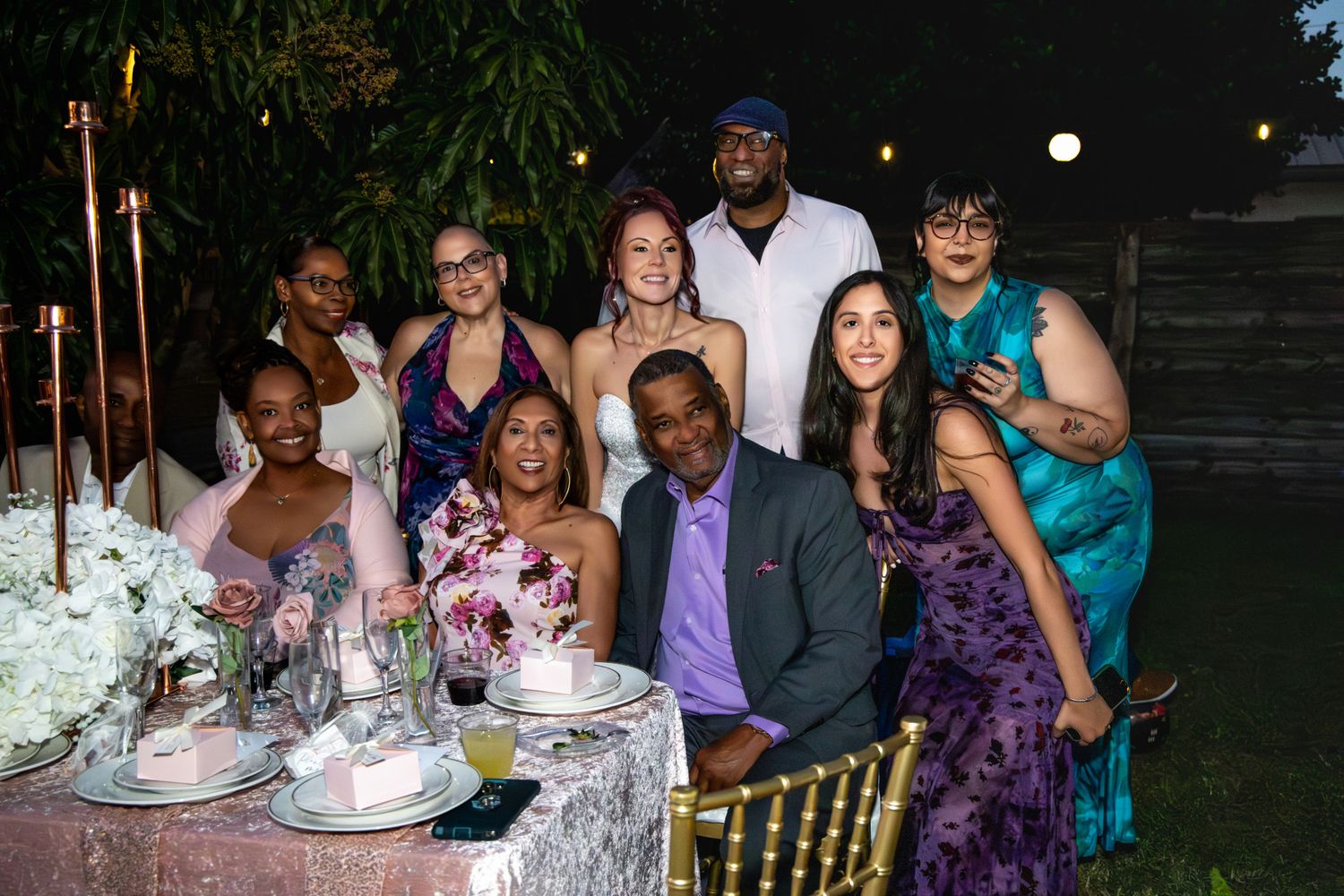 A group of formally dressed guests gather around an elegant table at an outdoor evening celebration.