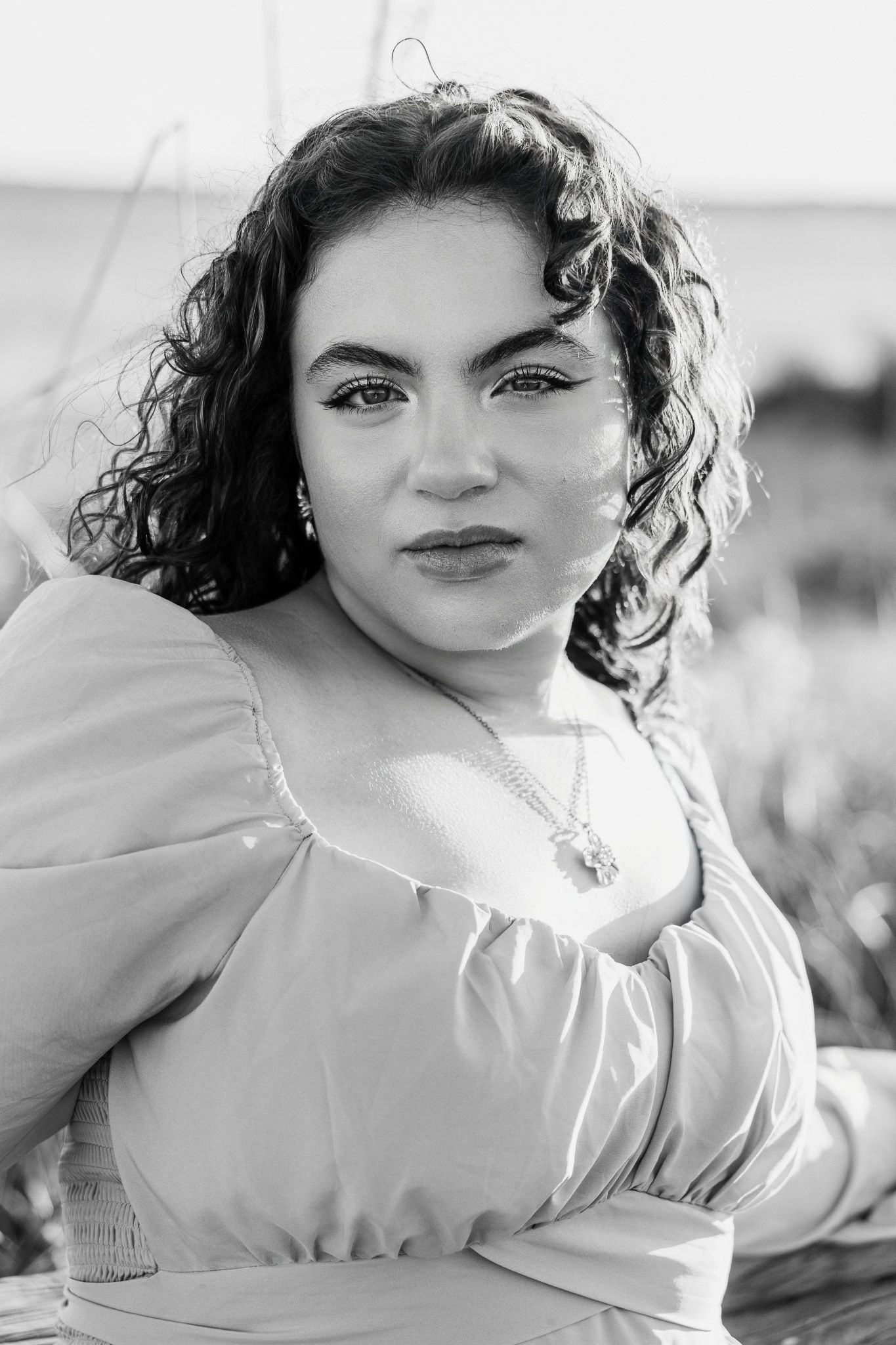 Black and white portrait of a young woman with curly dark hair wearing a puff-sleeve top and pendant necklace outdoors.