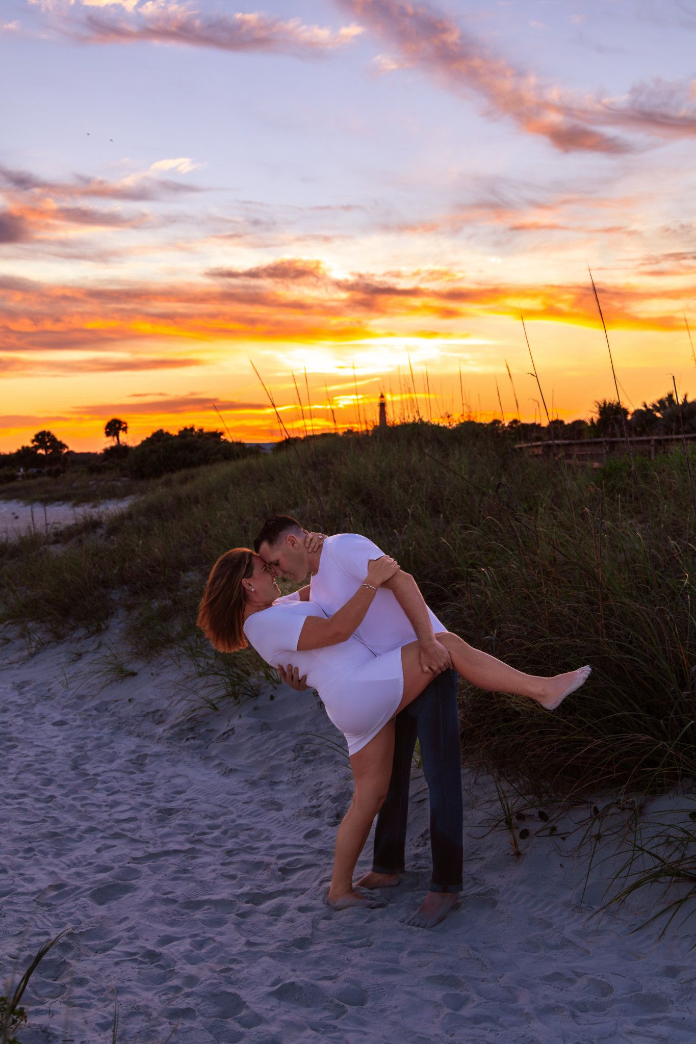 Couple sharing a romantic kiss at sunset on a sandy beach, with vibrant orange and purple skies behind them.
