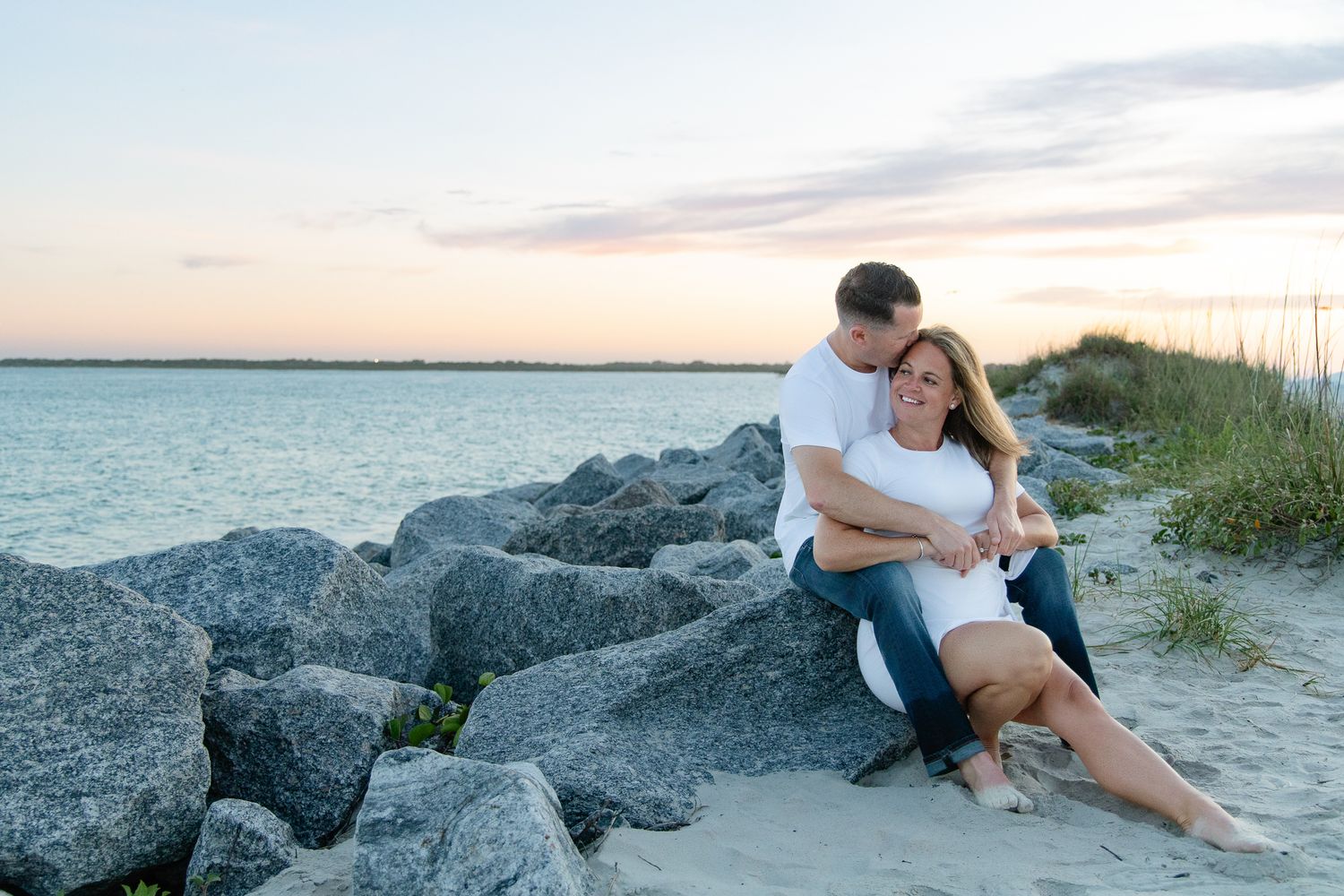 Couple sitting on rocky beach at sunset, man embracing woman in white top near calm ocean waters.