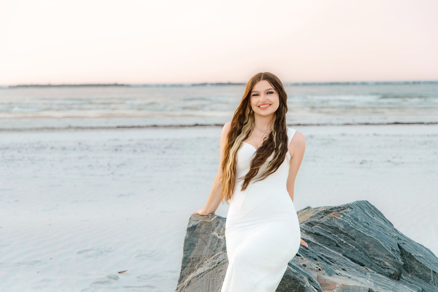Young woman in white dress smiling on rocky beach at sunset with calm ocean waves in background.