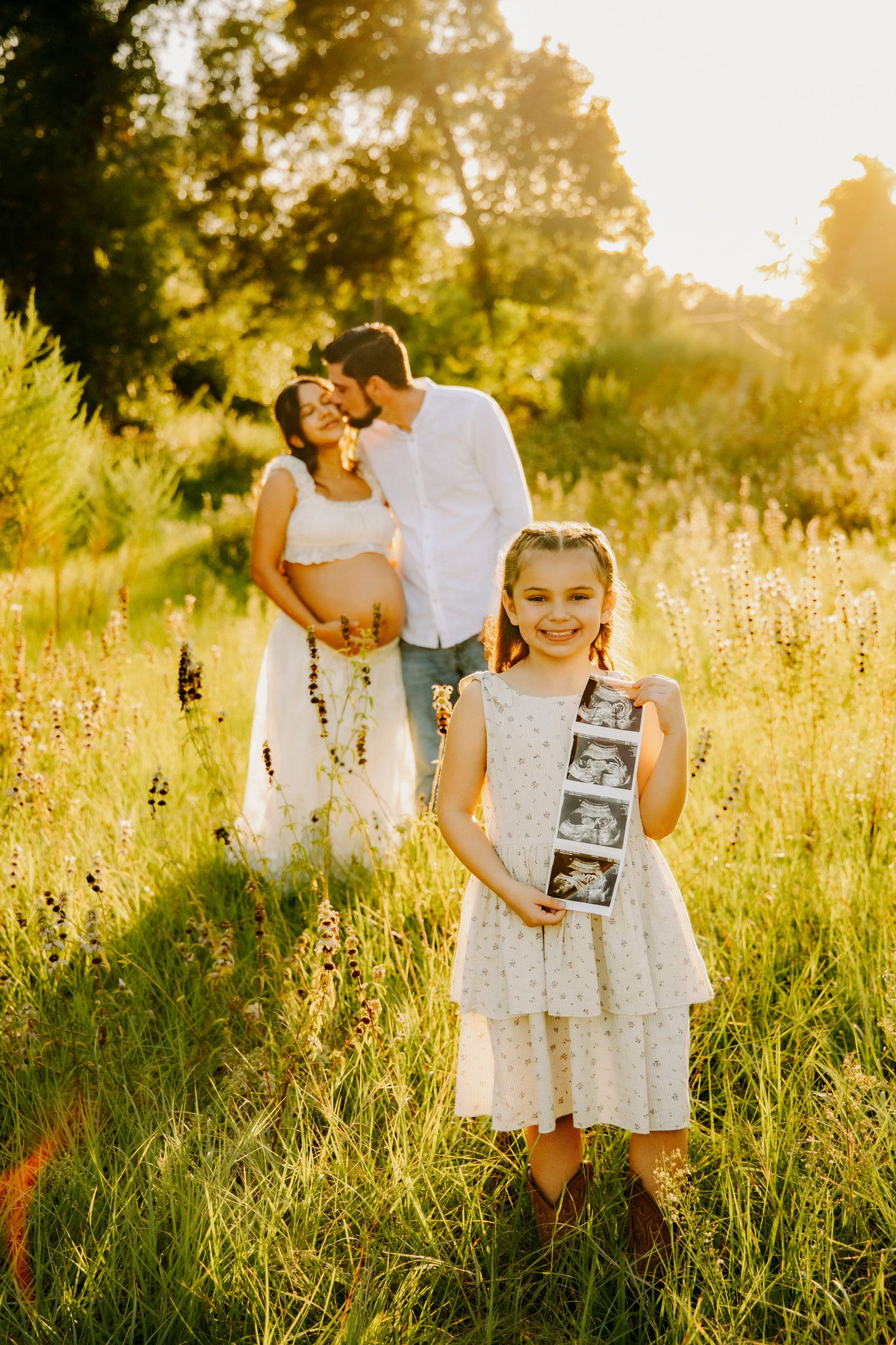 A young girl holds ultrasound photos in a sunny field while her pregnant mother and father share a kiss behind her.