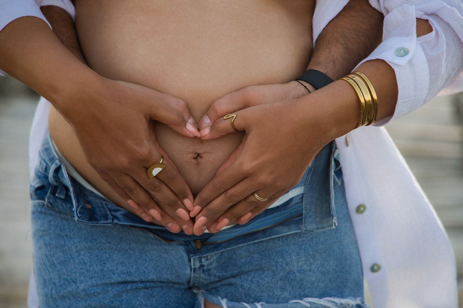 Couple forming heart shape with hands on pregnant belly, wearing gold jewelry and casual denim.