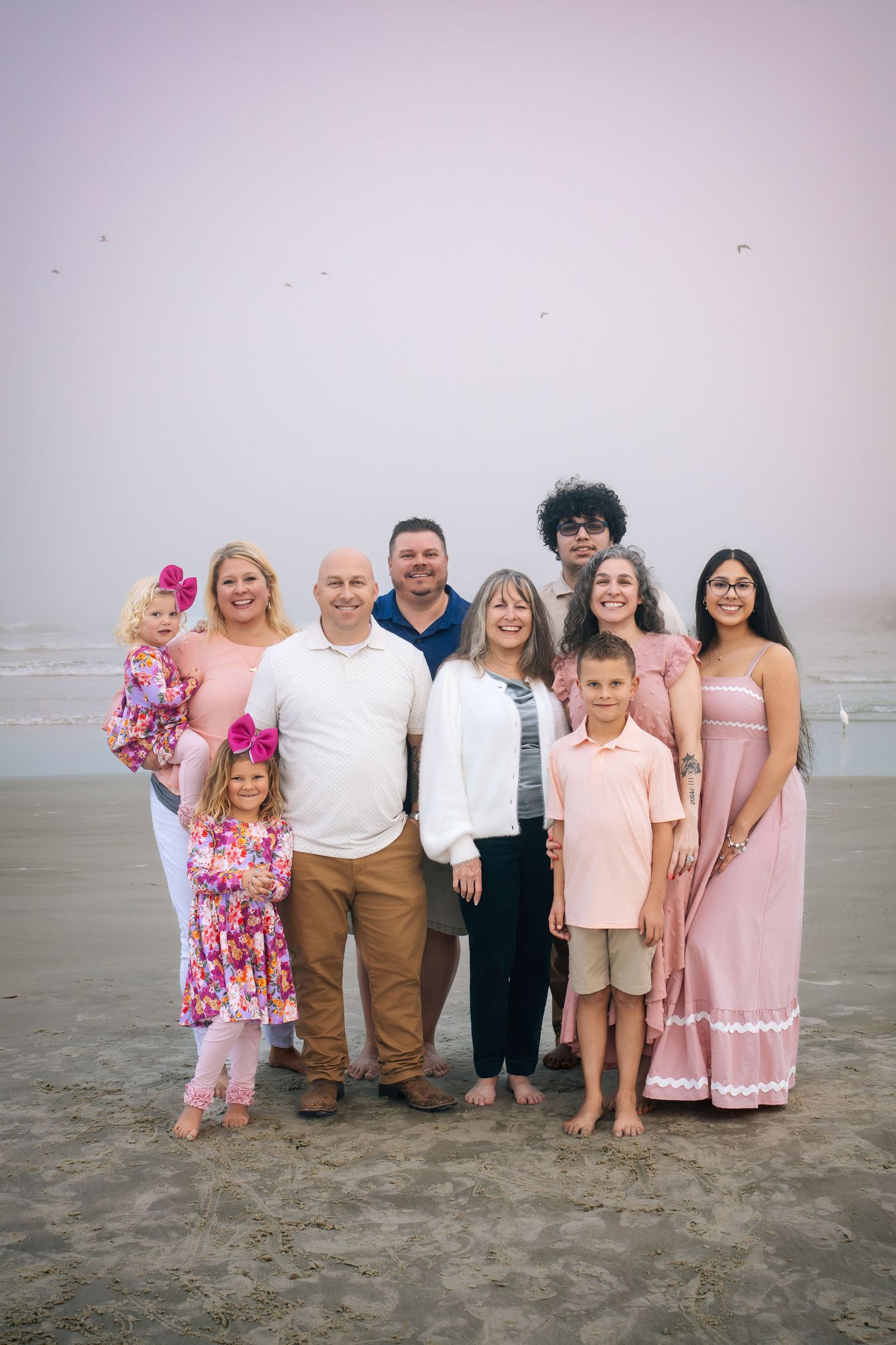 A family group in coordinated pink and neutral outfits poses together on a sandy beach at sunset.