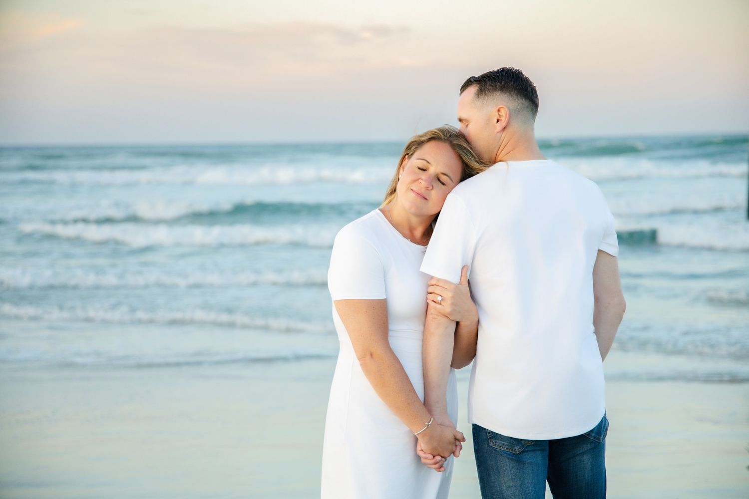Couple in white embracing on a beach at sunset, holding hands with ocean waves in the background.