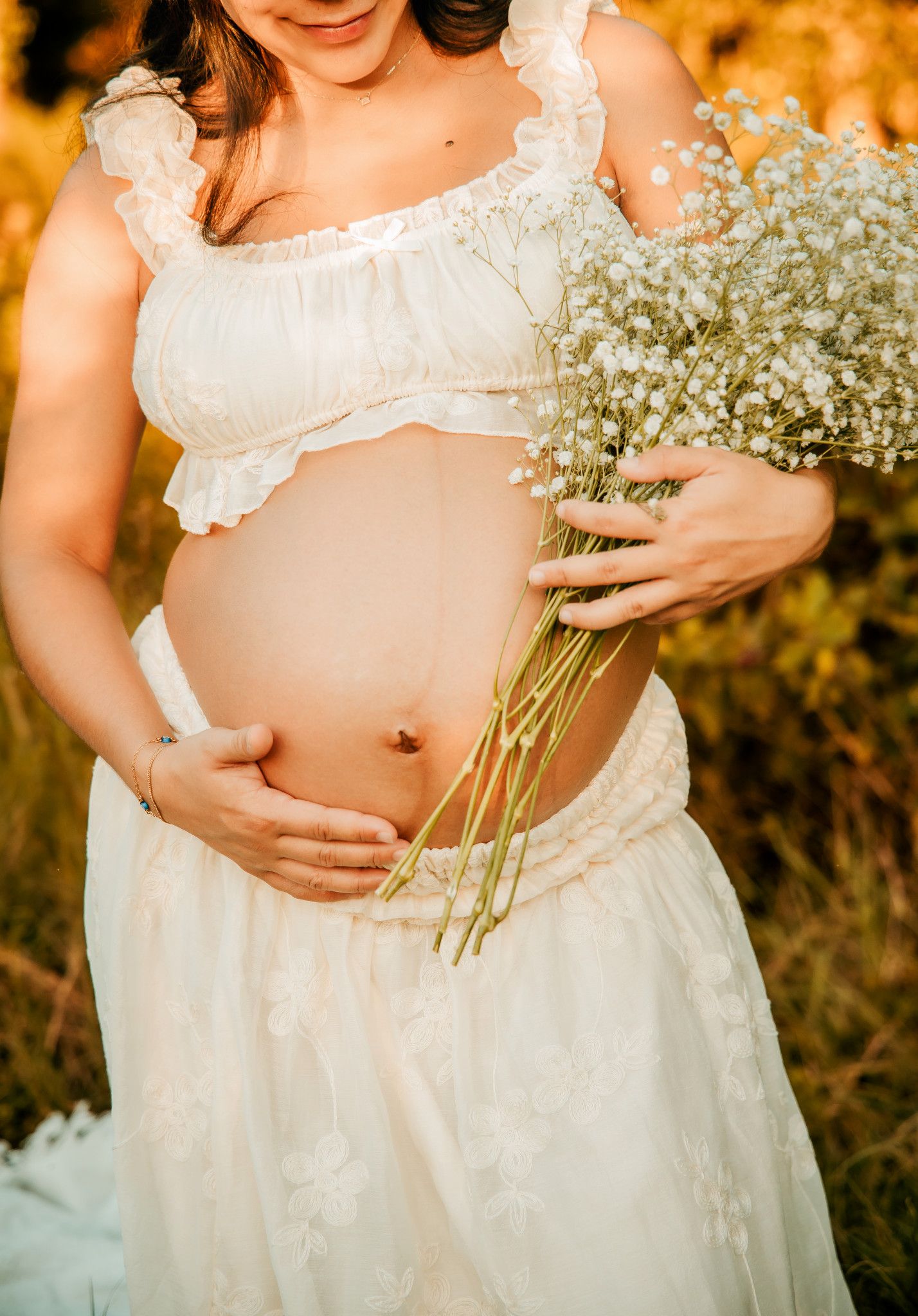 Pregnant woman in white crop top and skirt holding baby's breath flowers outdoors in golden sunlight.
