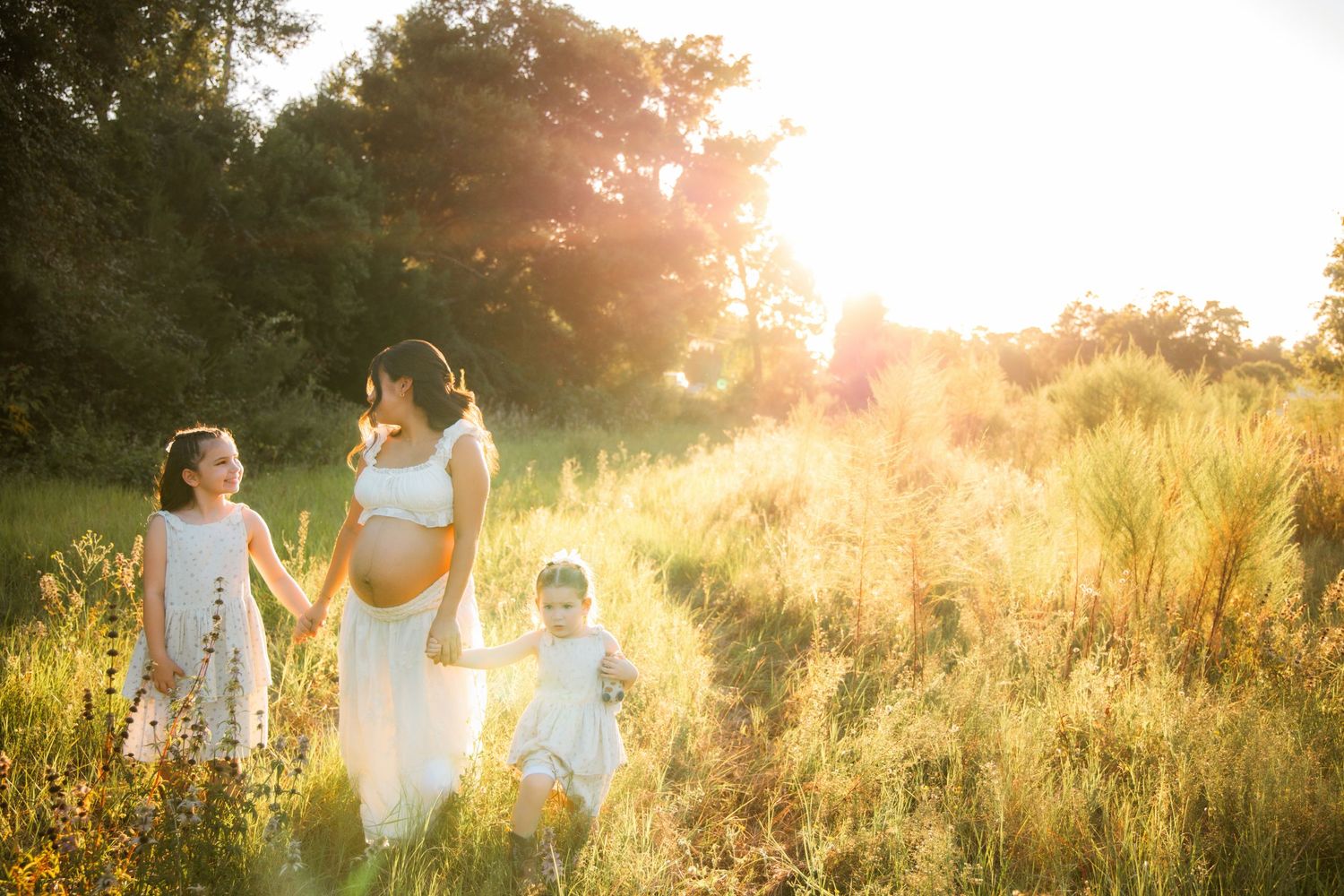 Pregnant woman walks through golden sunlit meadow holding hands with two young children in white dresses.