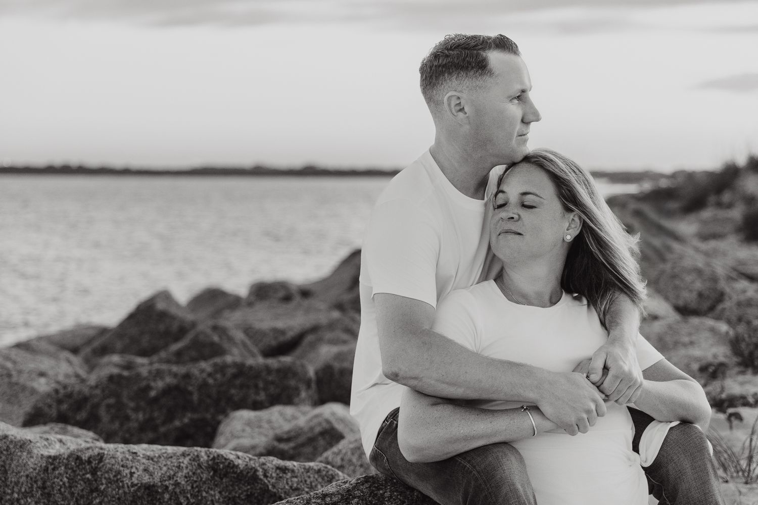 Couple embracing on rocky shore at sunset in black and white, romantic engagement photo by the water.