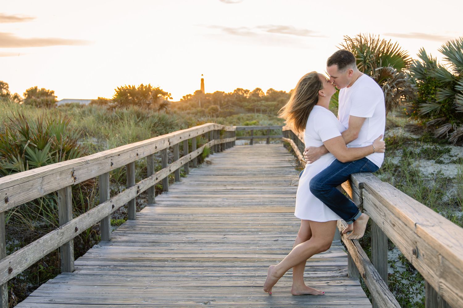 Couple sharing a romantic moment on a wooden boardwalk at sunset near a beach with lush greenery.
