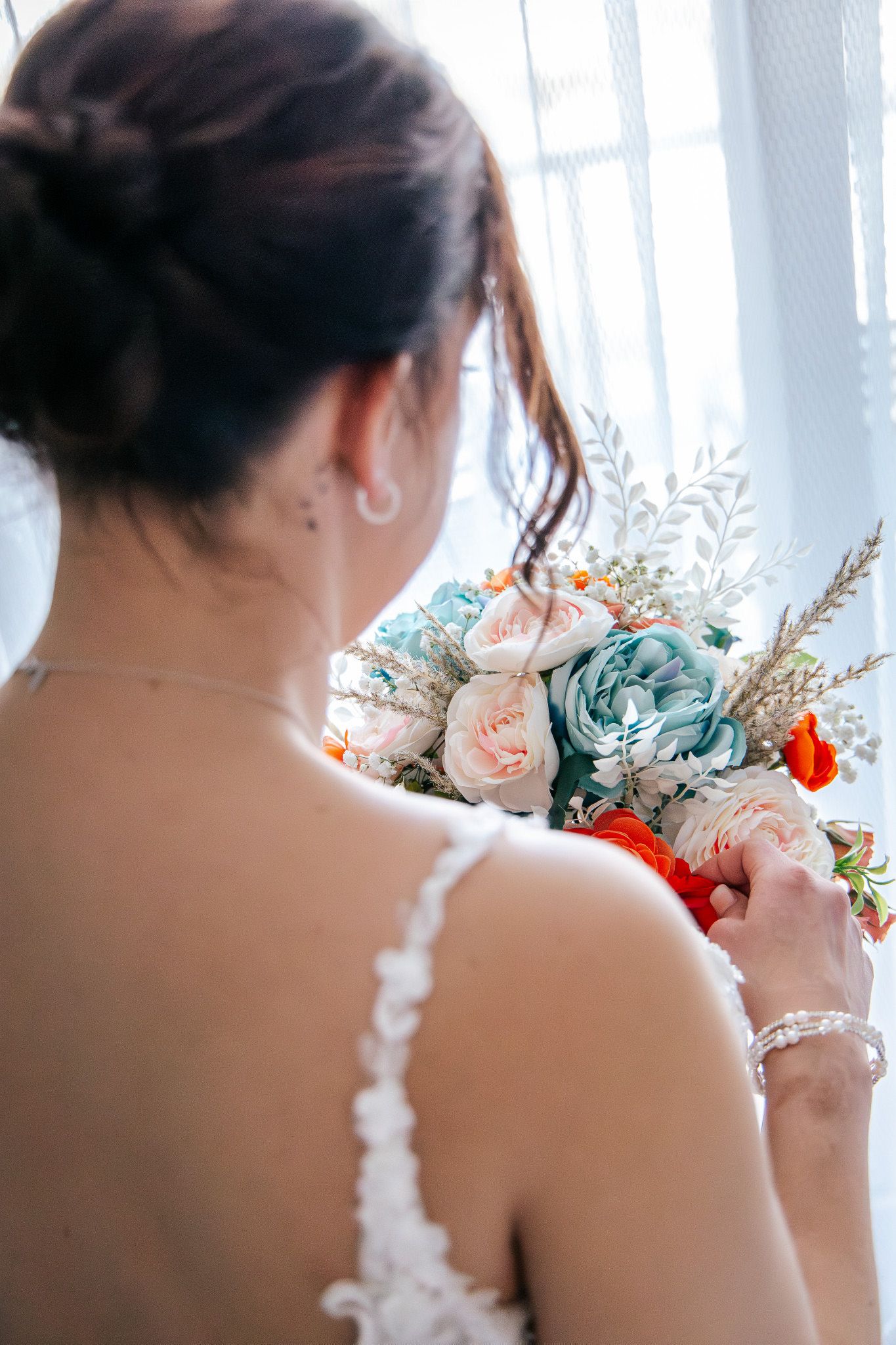 A bride in a white lace dress holds a beautiful bouquet of pink, white and orange flowers by a window.