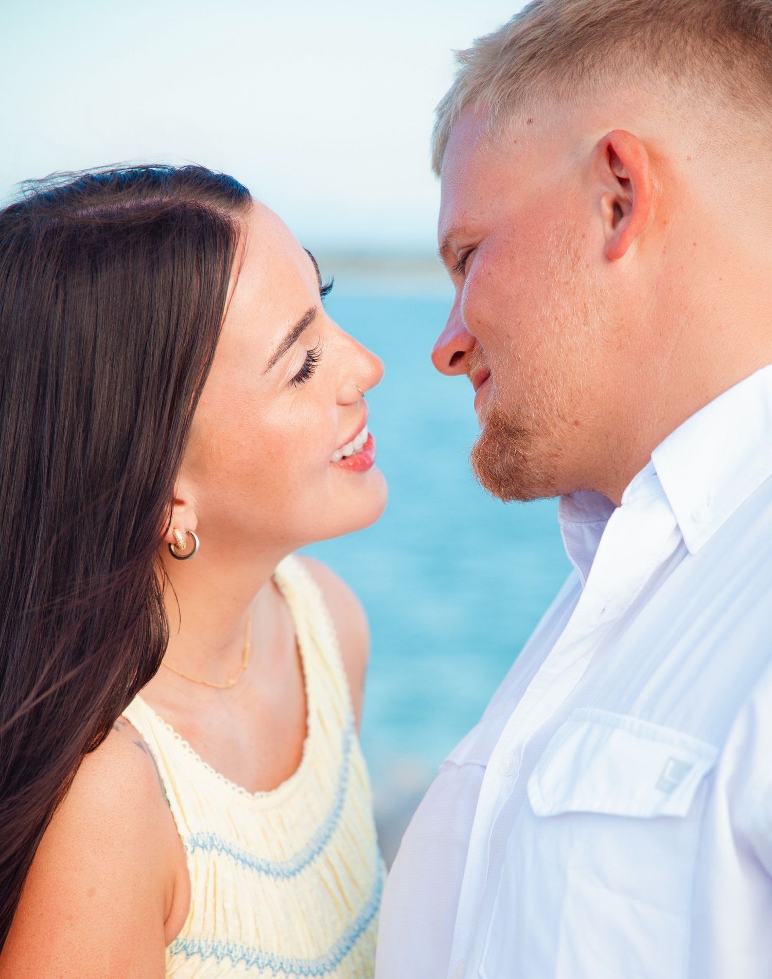 Close-up of smiling couple nearly kissing, woman in cream dress, man in white shirt, ocean background.