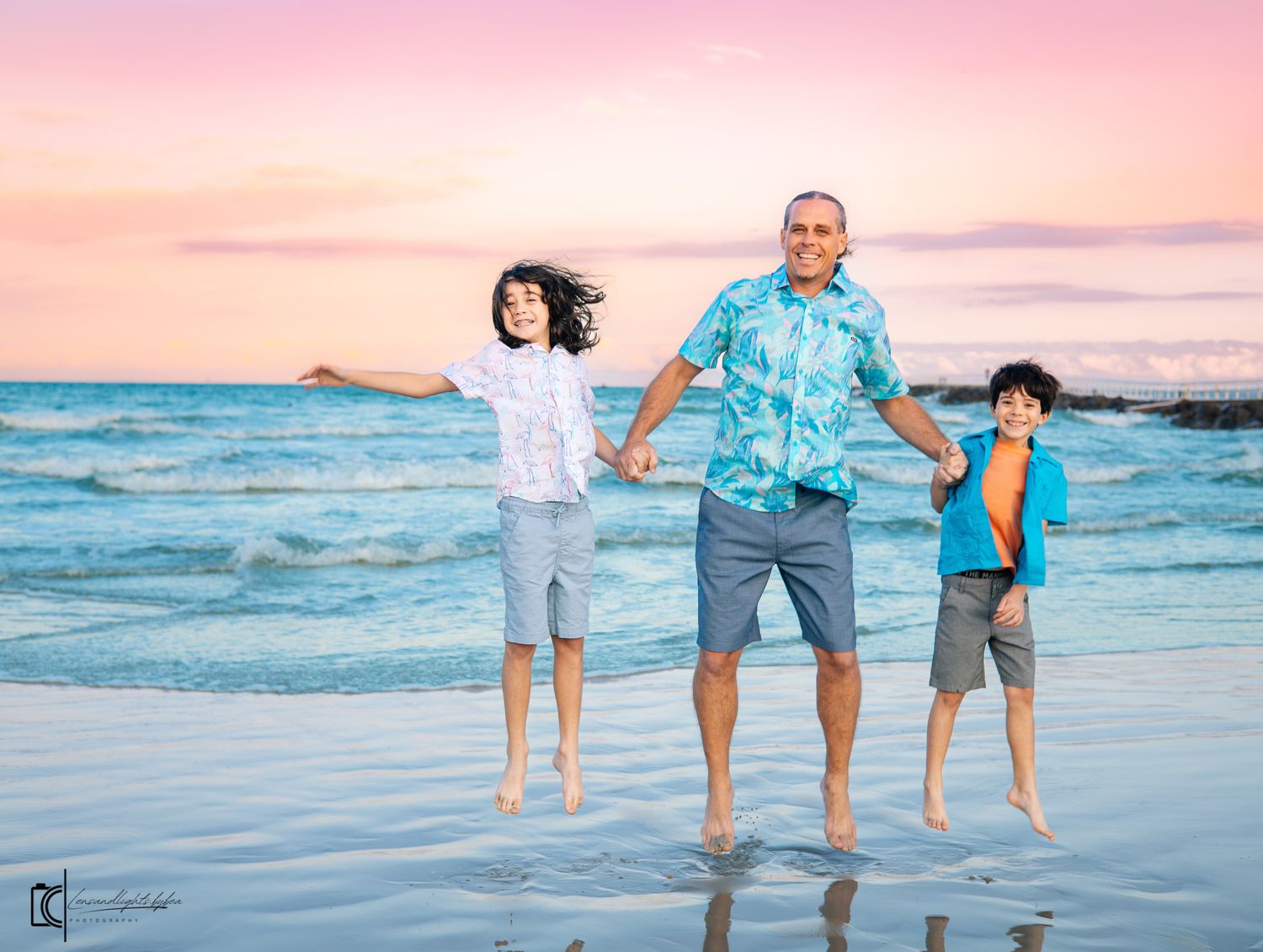 Family walking along beach shoreline at sunset with waves lapping at their feet in coordinated blue outfits.