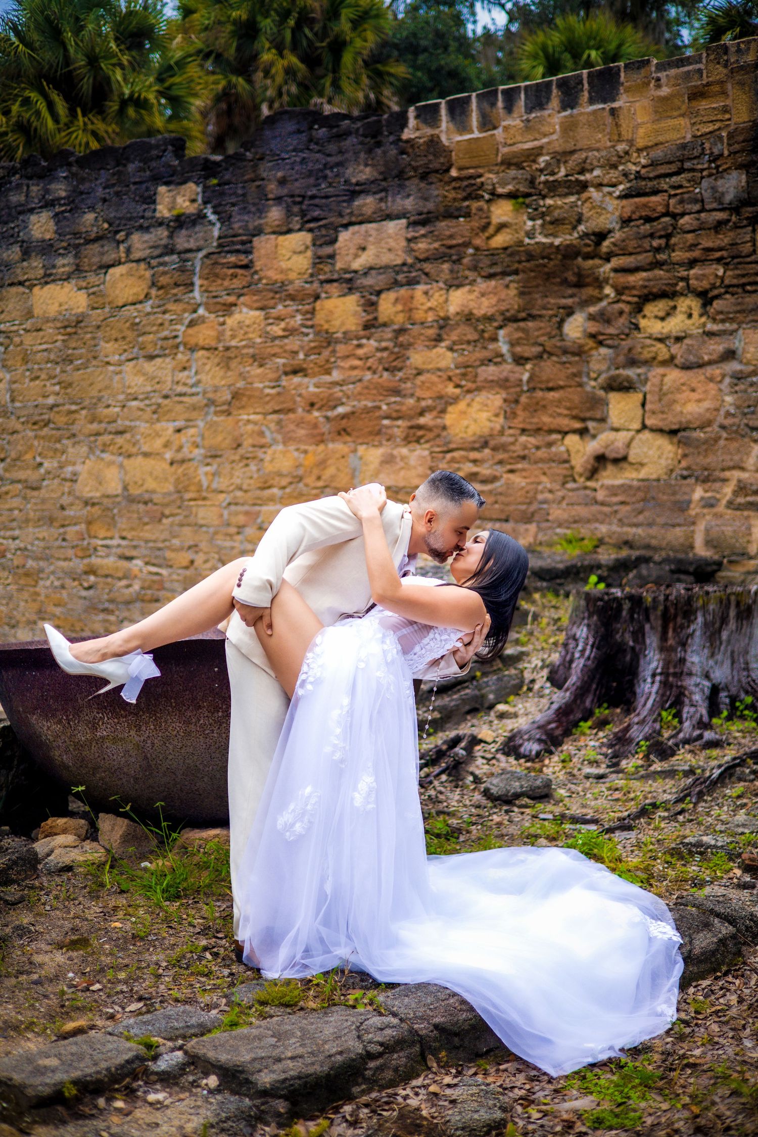 Playful wedding couple poses near historic stone wall in outdoor garden setting.