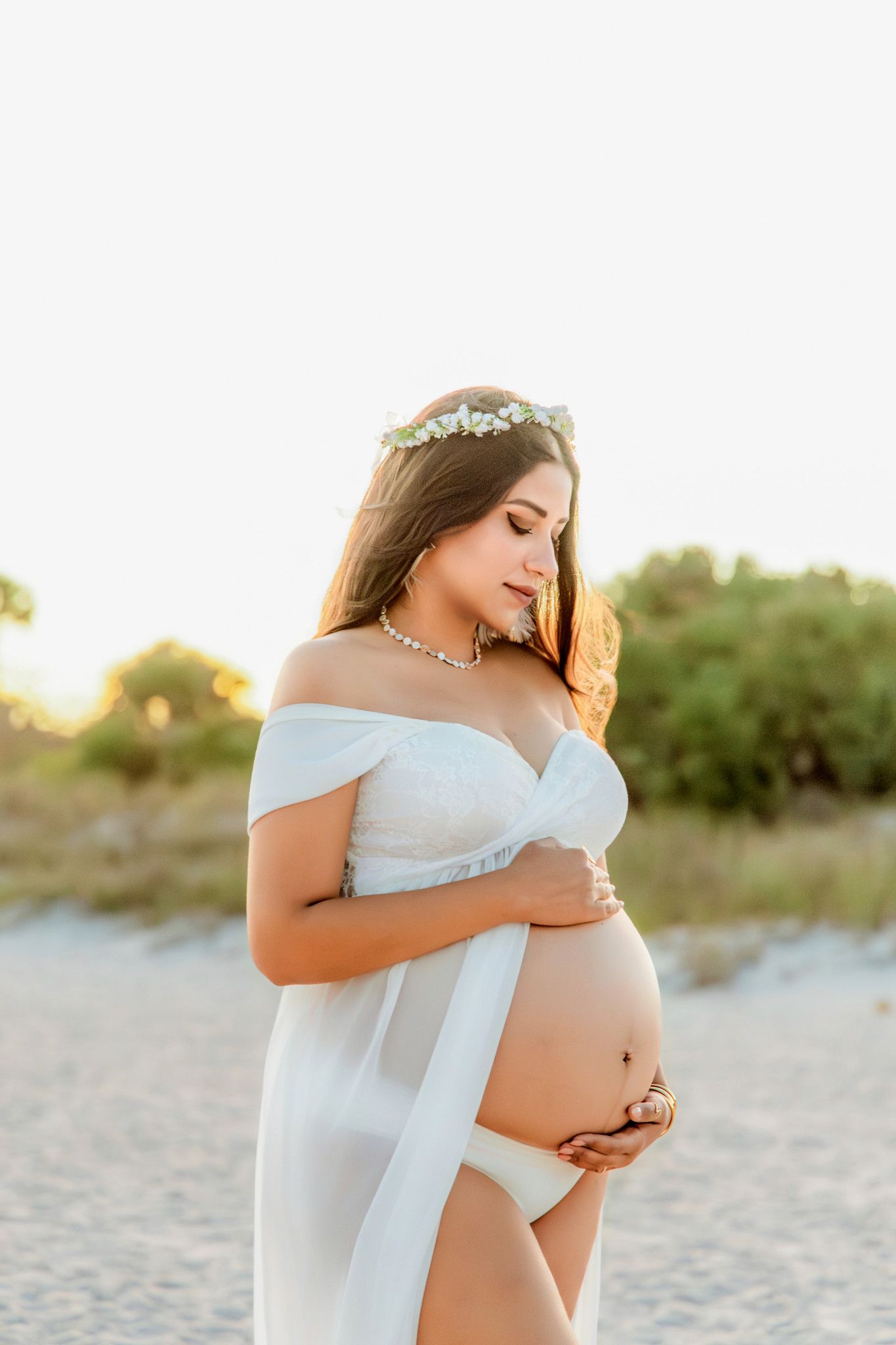 Pregnant woman in white off-shoulder dress with floral crown poses on beach at golden hour, cradling her baby bump.