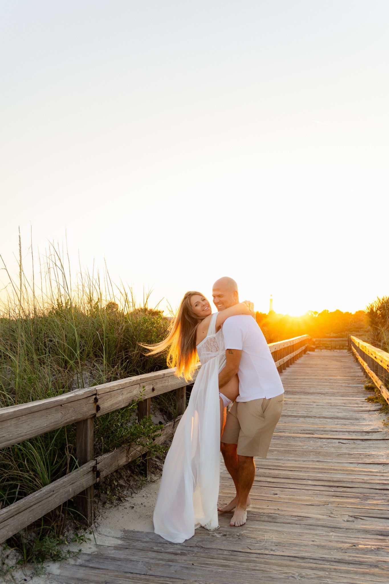 Couple embracing on a wooden boardwalk at golden sunset, woman in white flowing dress, man in white shirt and khaki shorts.