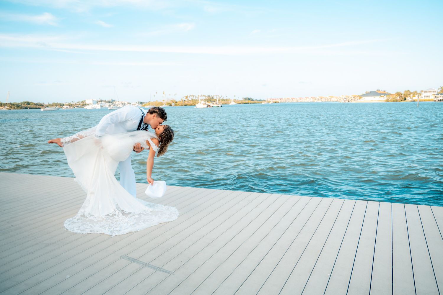 Bride and groom share a romantic dip kiss on a waterfront dock with scenic ocean views on their wedding day.