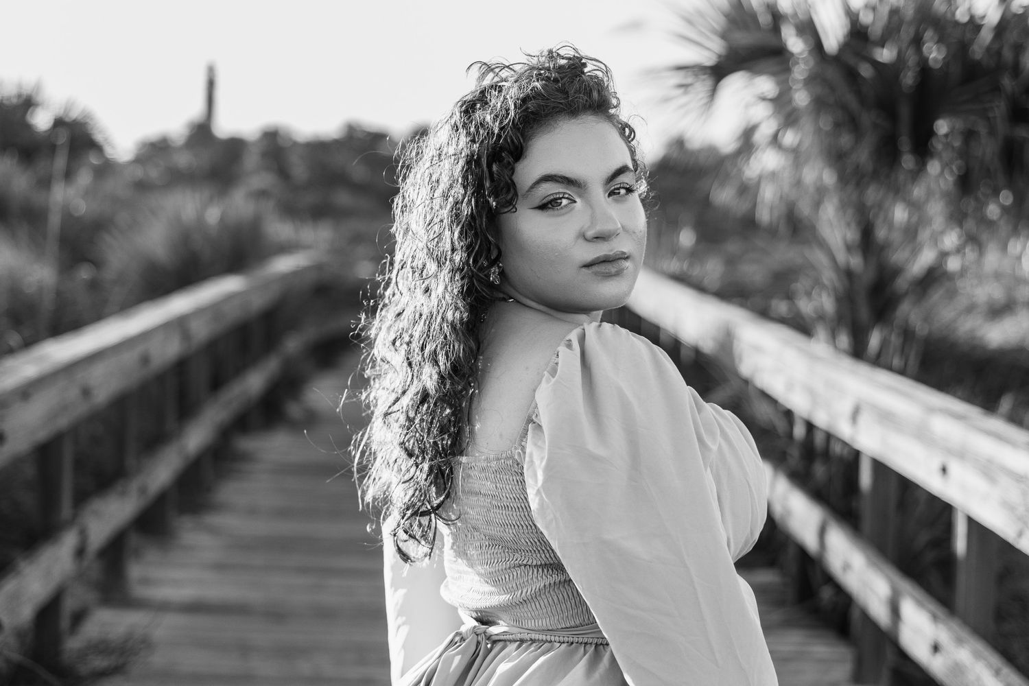 Black and white portrait of a young woman with long curly hair looking over her shoulder on a wooden boardwalk with palm trees behind her.