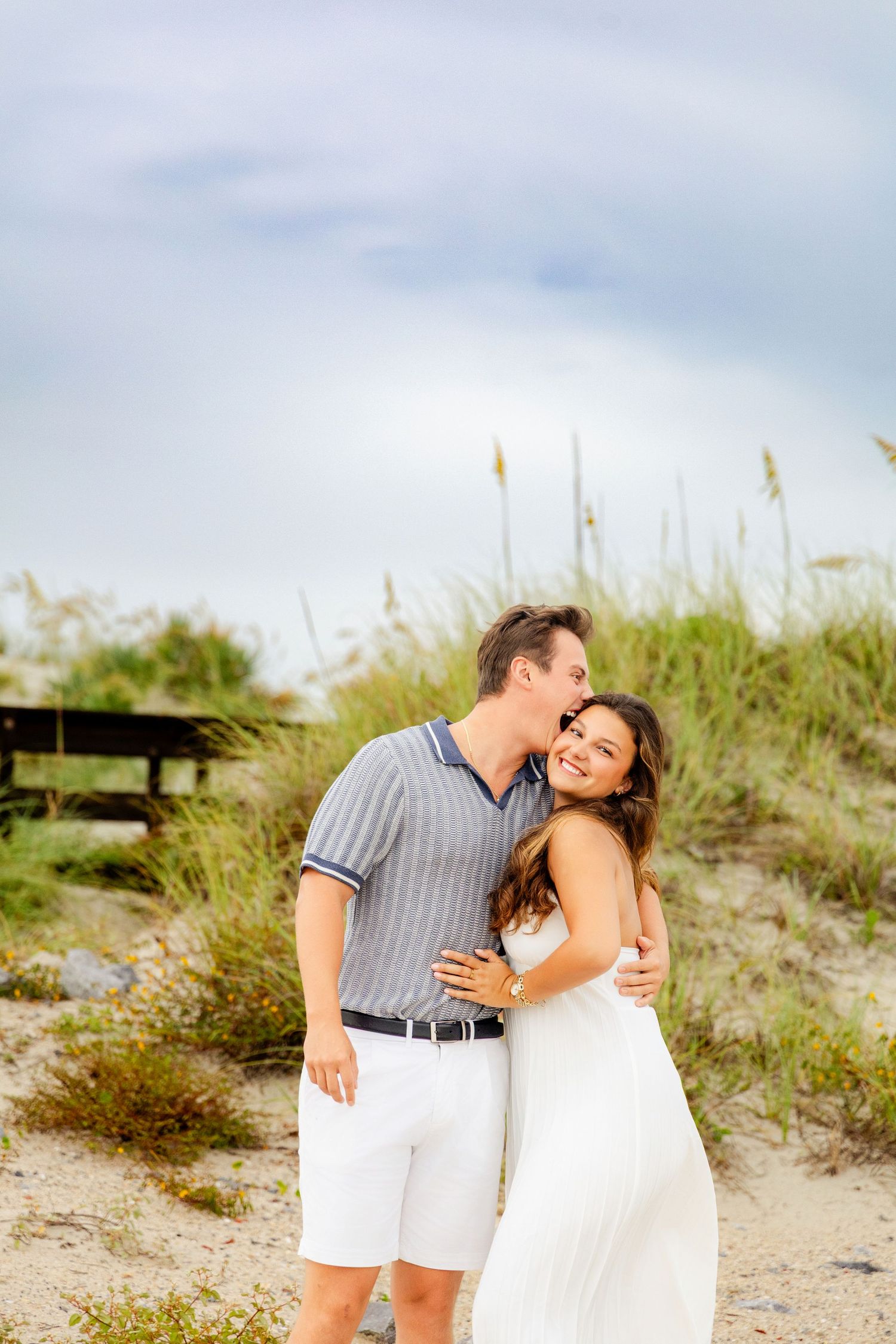 Couple embracing on a sandy beach, man kissing woman's cheek, she smiles in white dress, dune grass background.
