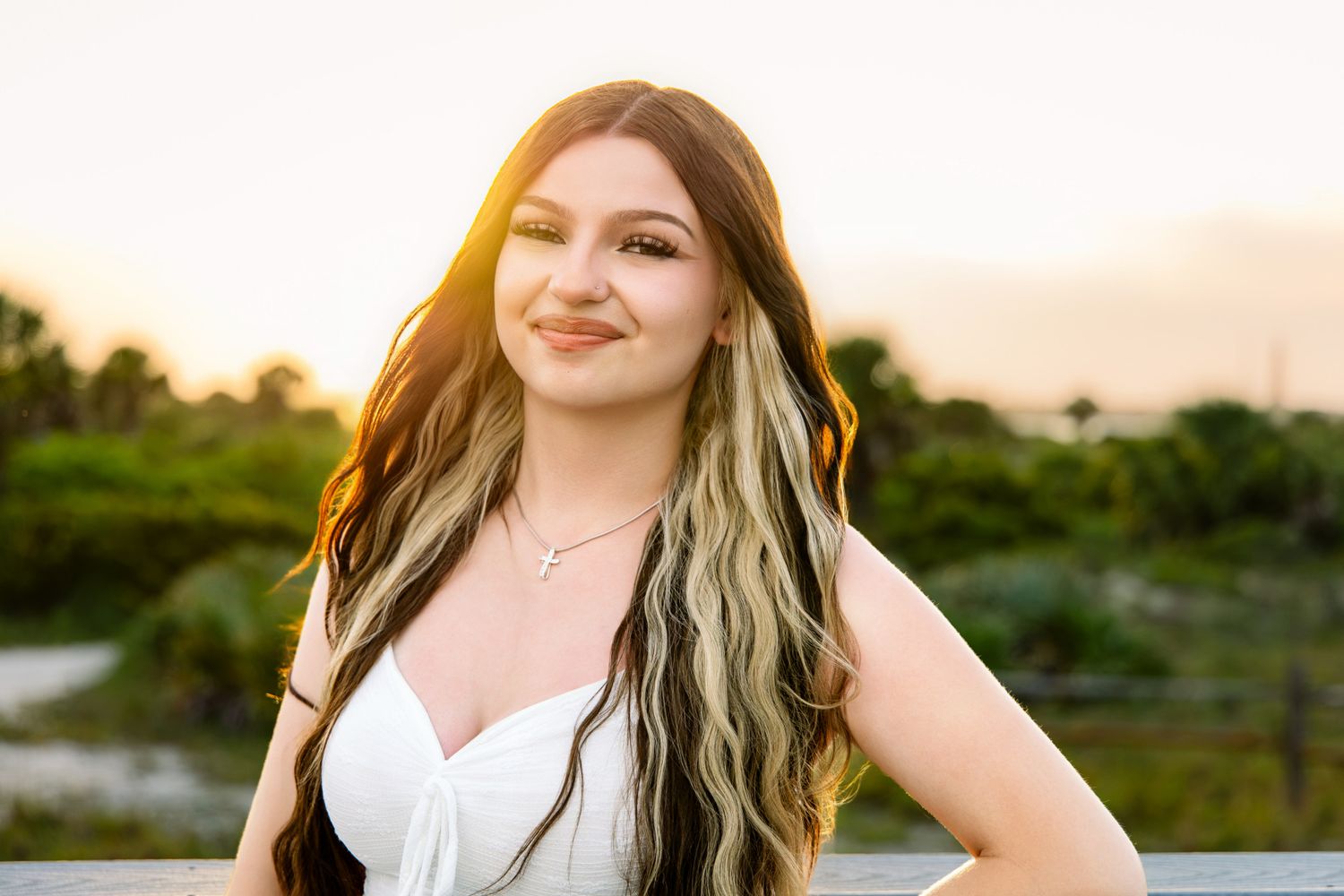 Young woman with long highlighted hair smiling at sunset on outdoor boardwalk.
