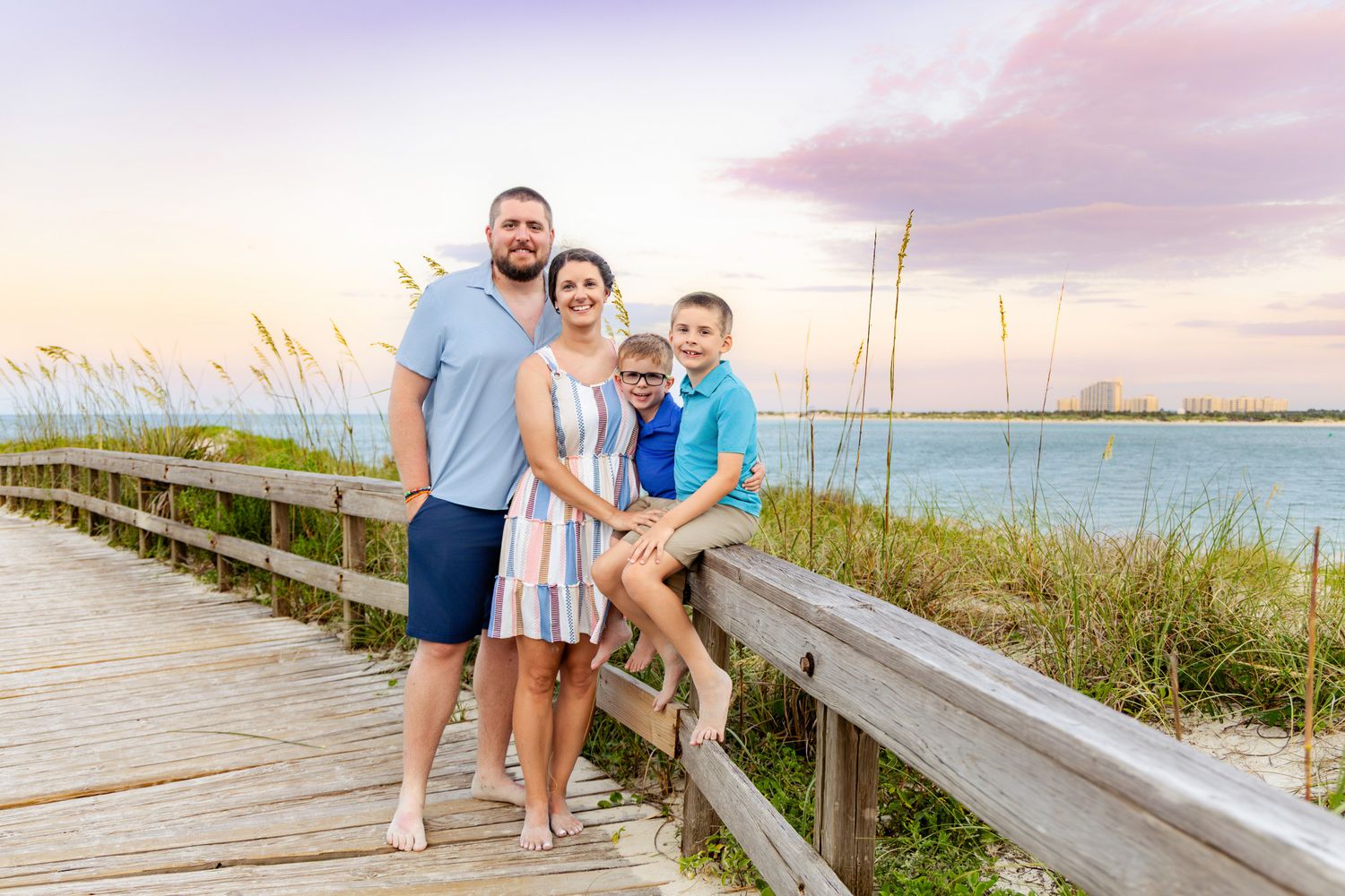 Family of four posing on a wooden beach boardwalk at sunset with sea oats and ocean views.