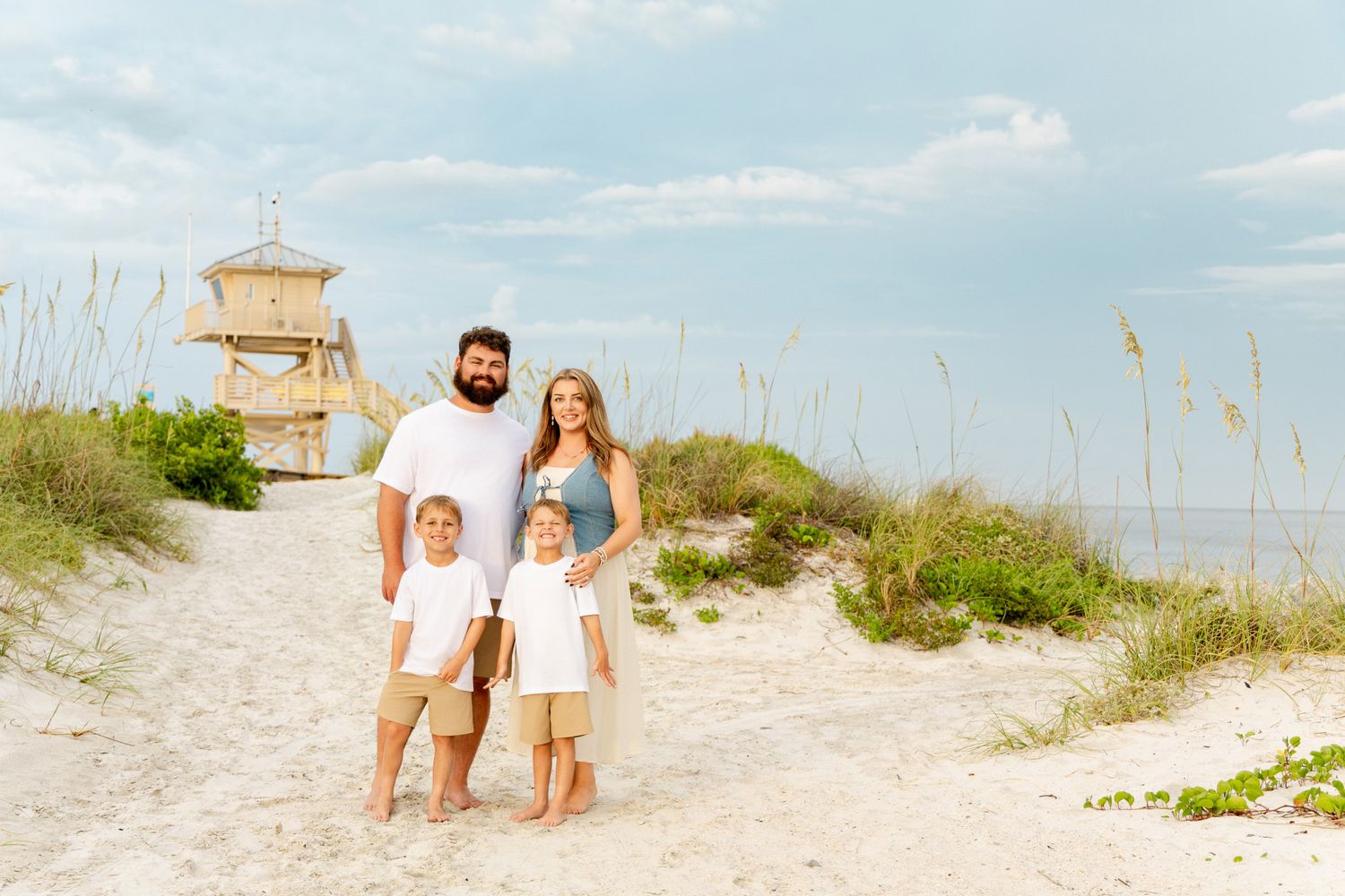 Family of four poses on sandy beach path with sea oats and lifeguard tower, smiling under blue skies.