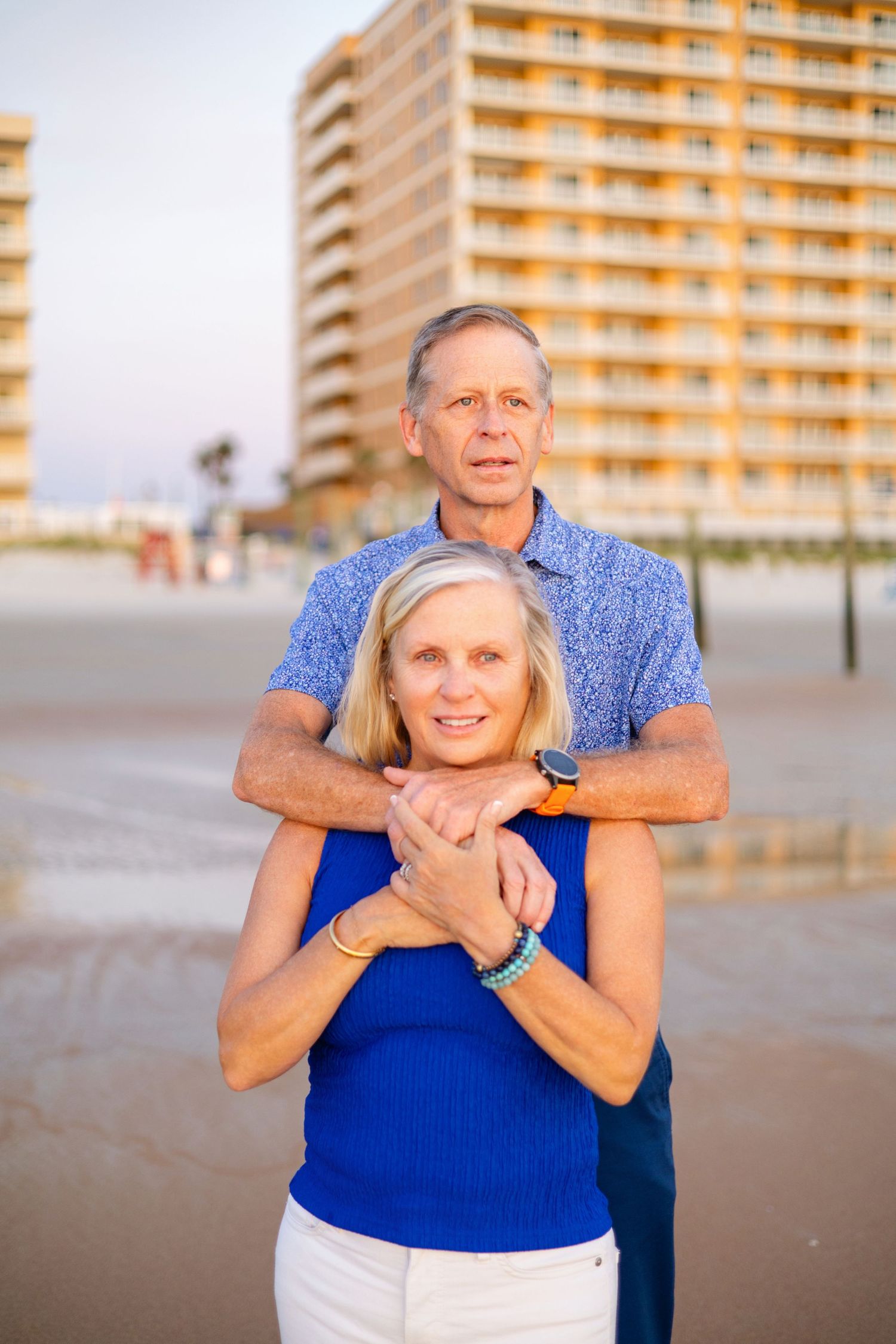 A senior couple in matching blue outfits embrace on a beach with a hotel building in the background.