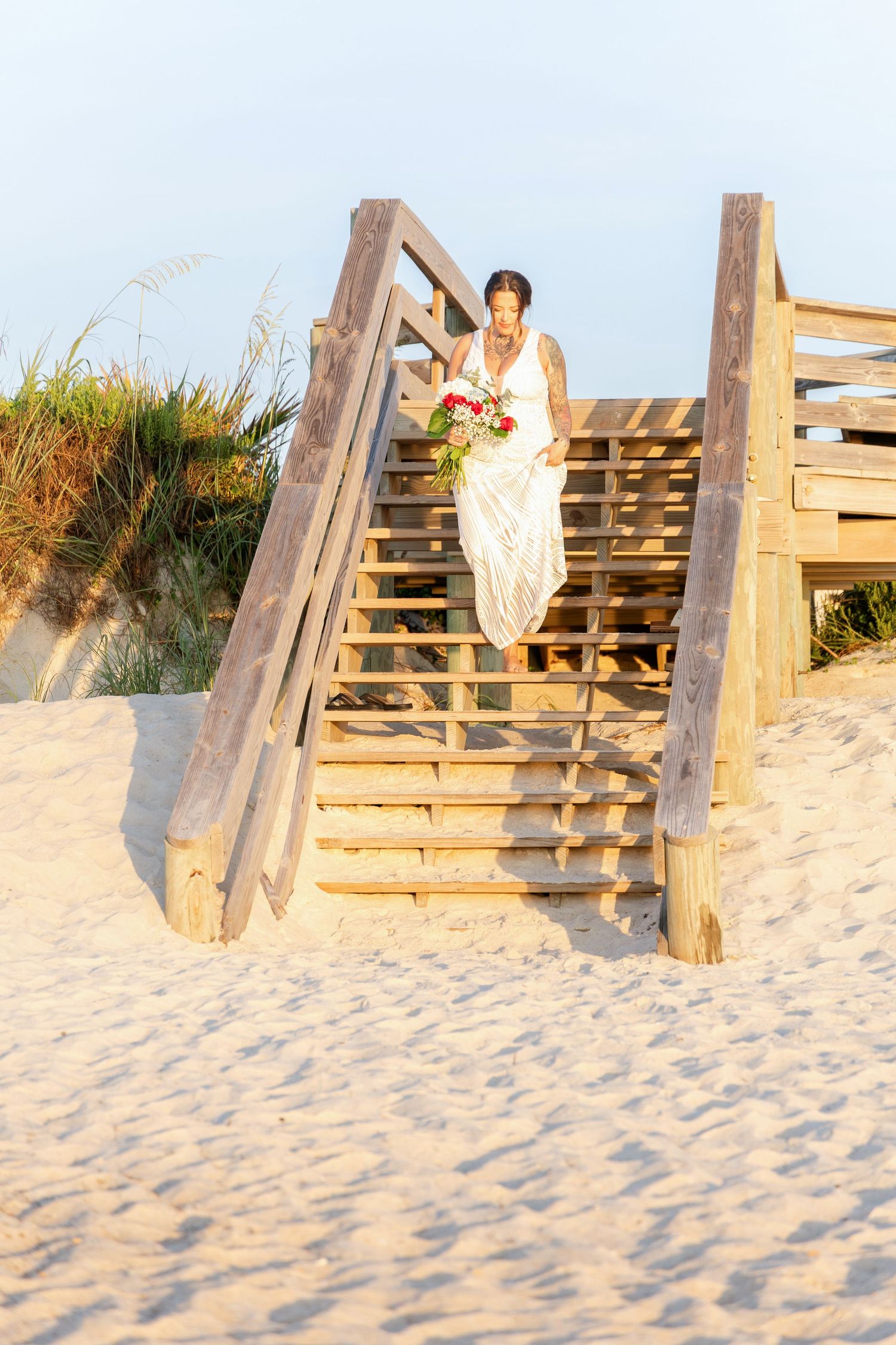 Tattooed bride in white lace gown descending wooden beach boardwalk stairs holding bridal bouquet.
