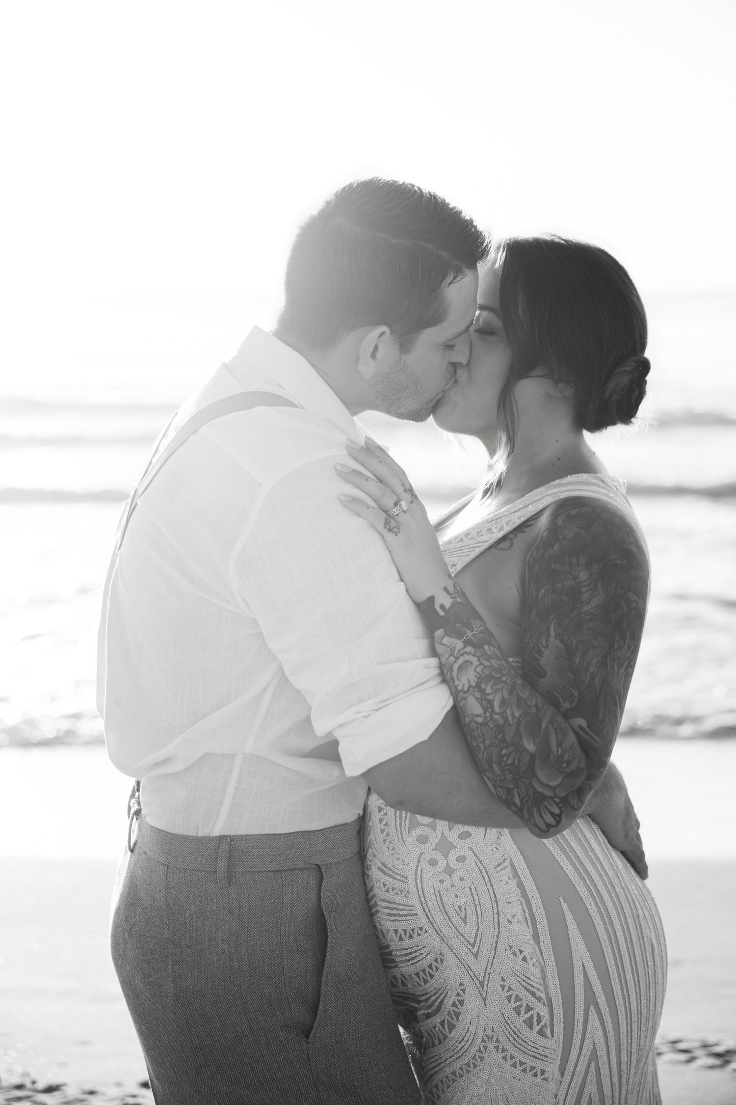 Black and white photo of a couple sharing a romantic kiss on the beach at sunset, woman with tattoos in lace dress.