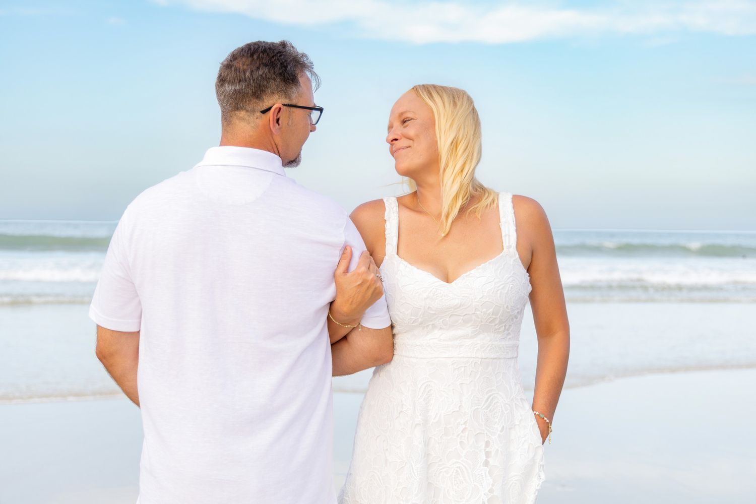 Couple in white outfits sharing a romantic moment on a sunny beach with ocean waves in the background.