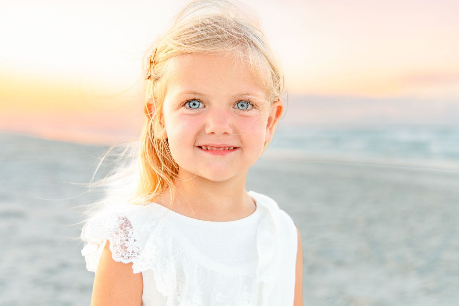 Adorable blonde girl with blue eyes smiling on the beach at sunset in a white dress.
