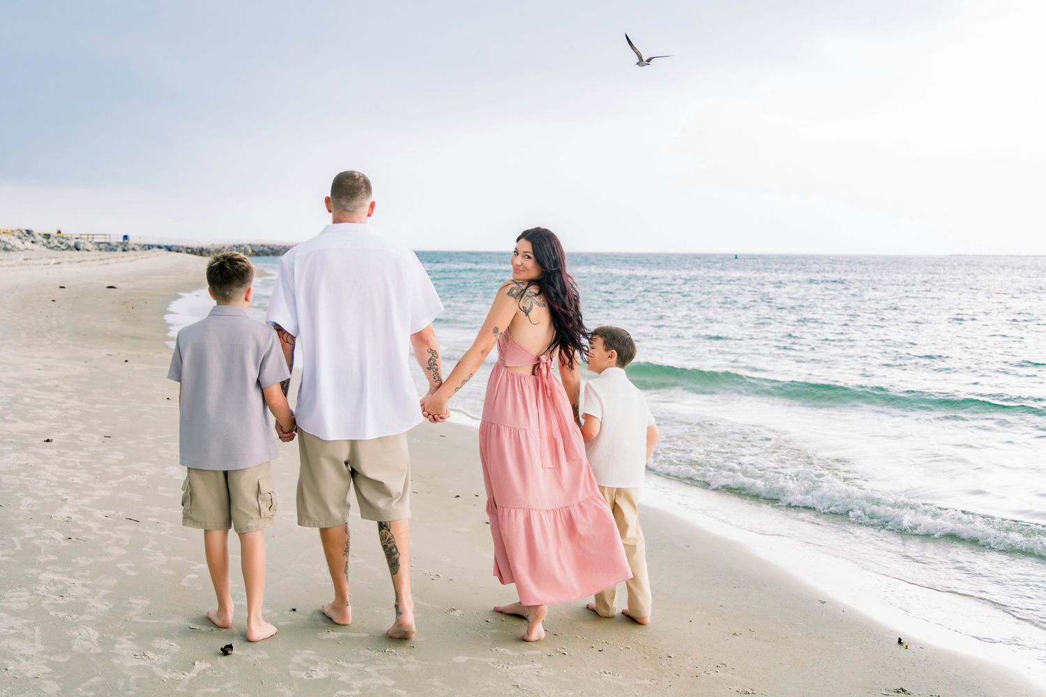 Family of four walking hand-in-hand along a sandy beach with ocean waves and a seagull flying overhead.