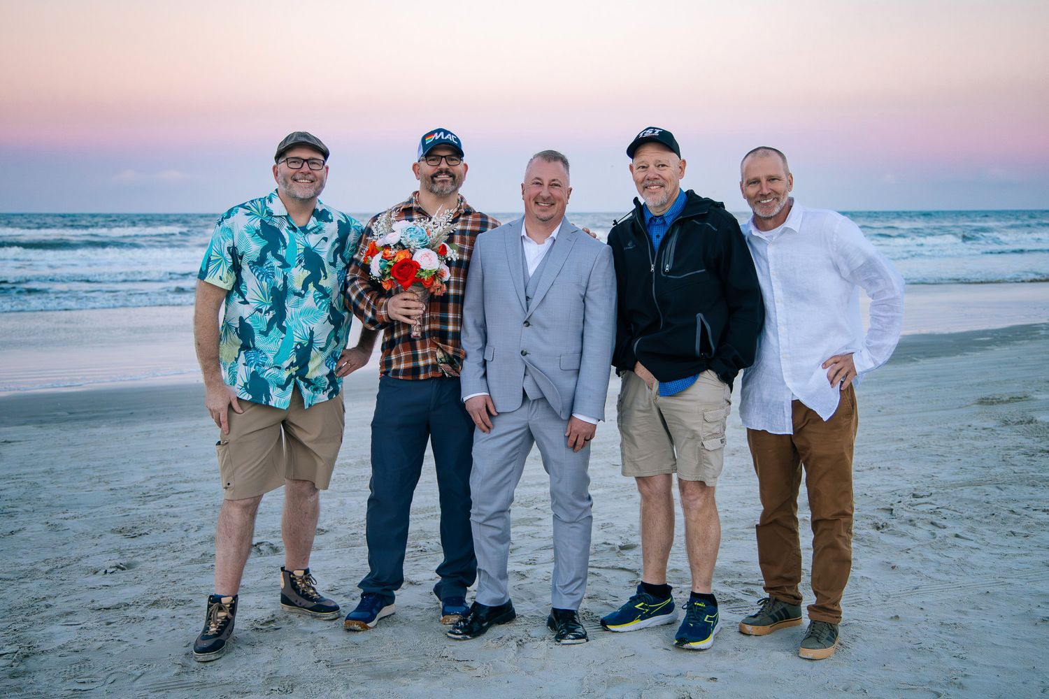 Group of people in casual attire standing together on a sandy beach at sunset.