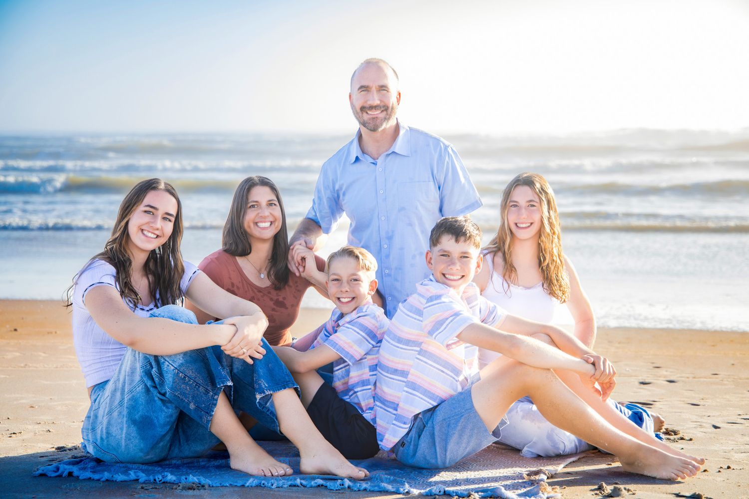 Family members sit casually on the sand at the beach during golden hour with waves rolling in behind them.
