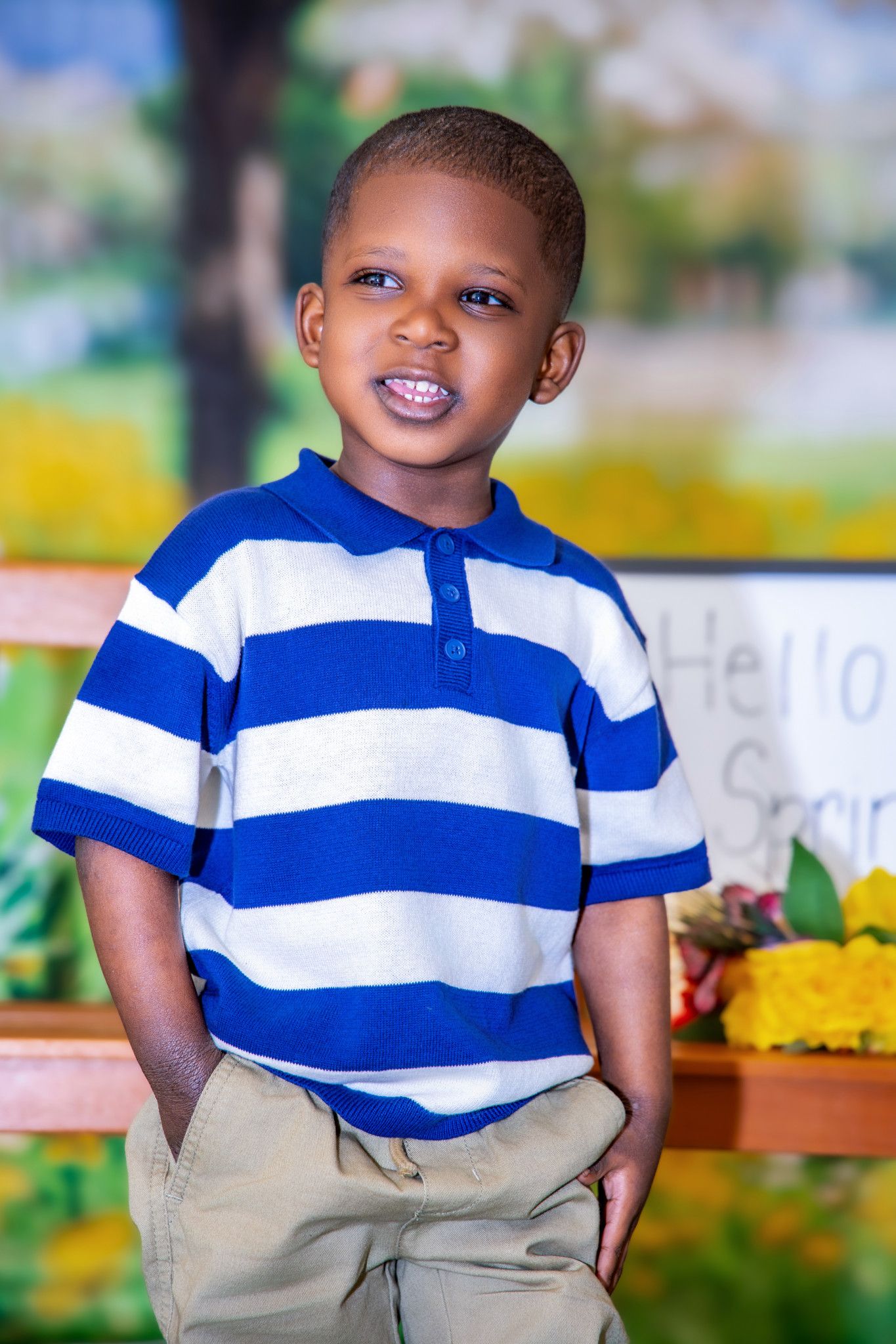 A young child in a blue and white striped polo shirt and khaki pants smiles outdoors in a natural setting.
