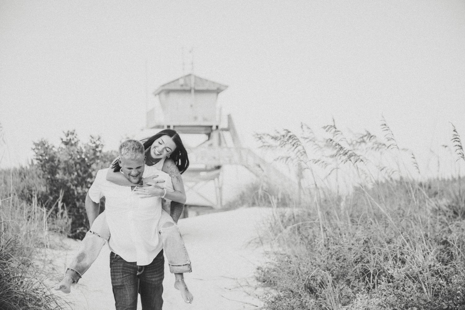 Black and white photo of couple laughing on beach path, man giving woman piggyback near lifeguard tower.