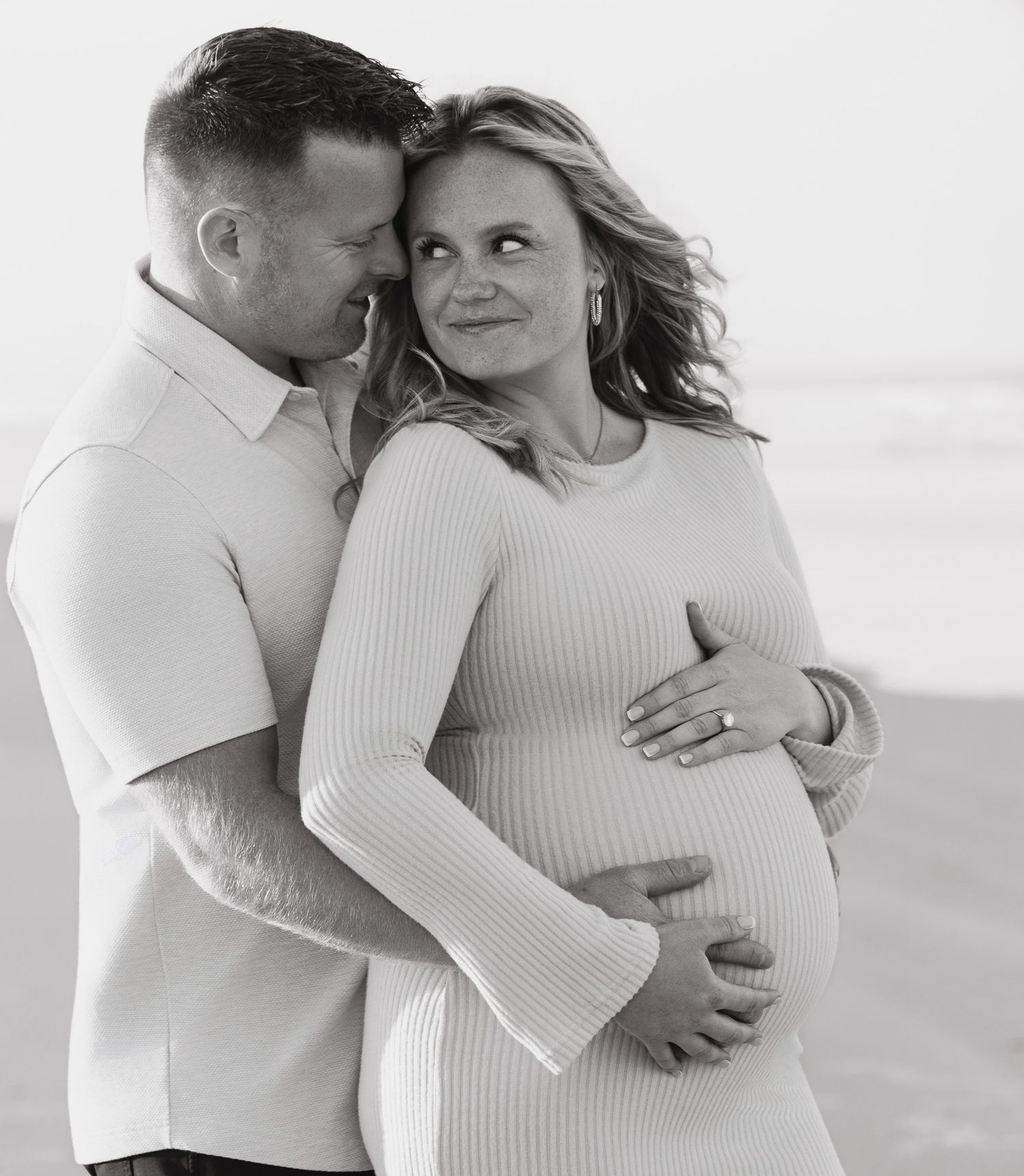 A romantic black and white maternity portrait of a couple embracing on the beach.