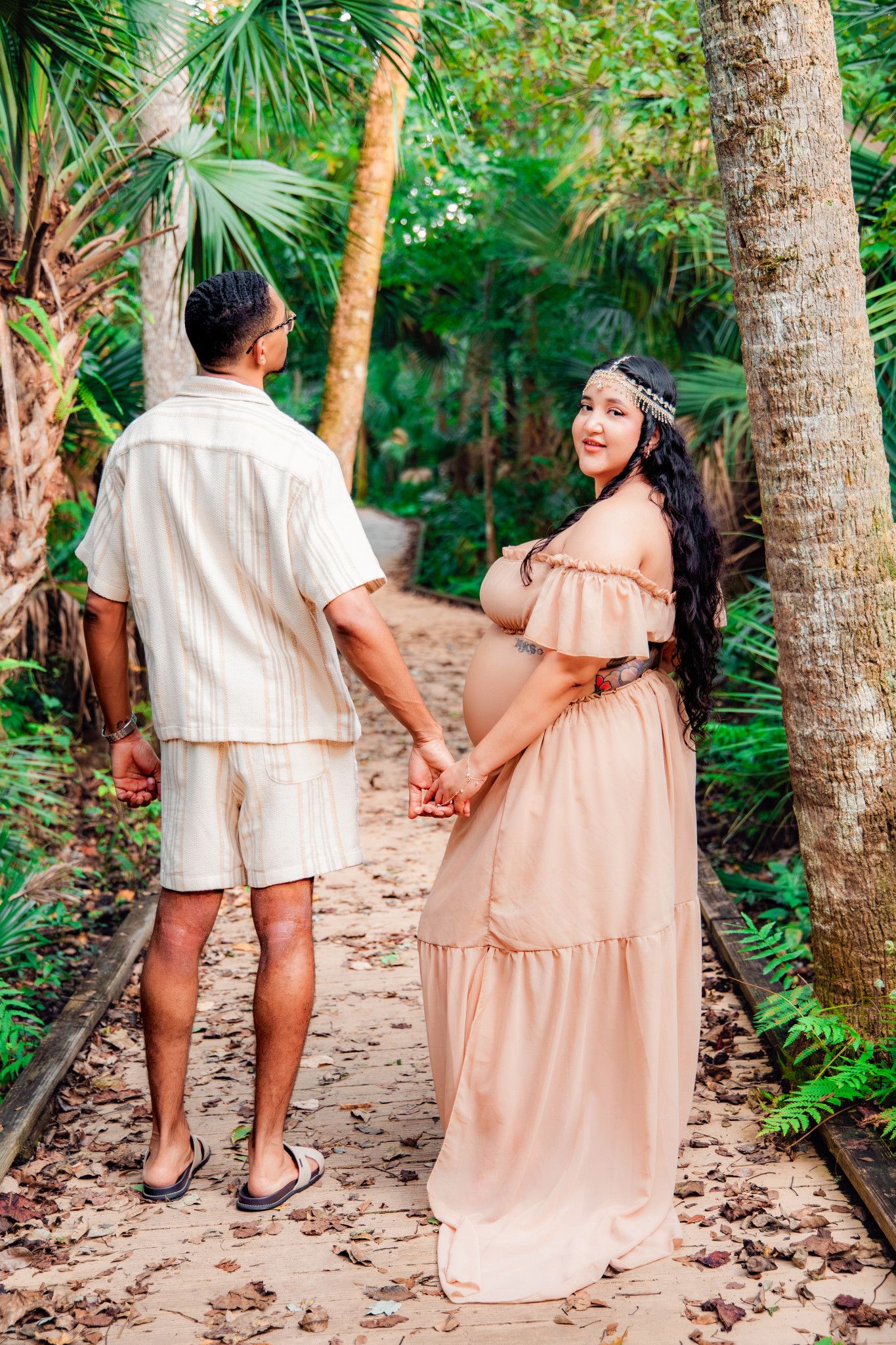 Pregnant woman in pink dress holds hands with man on tropical garden path surrounded by lush palm trees.