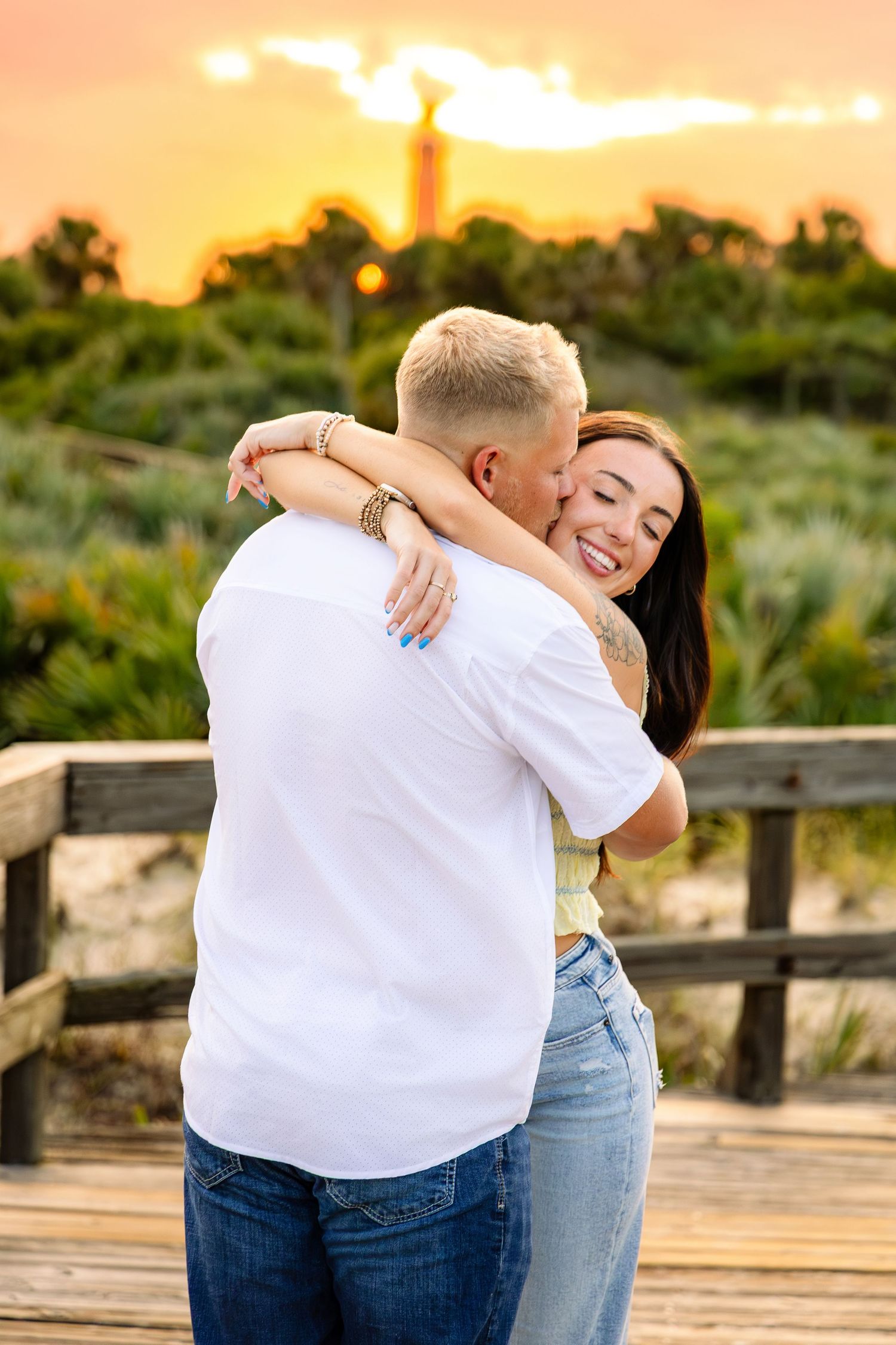 Couple embracing at sunset on a wooden boardwalk, woman smiling joyfully, with golden sky and greenery backdrop.