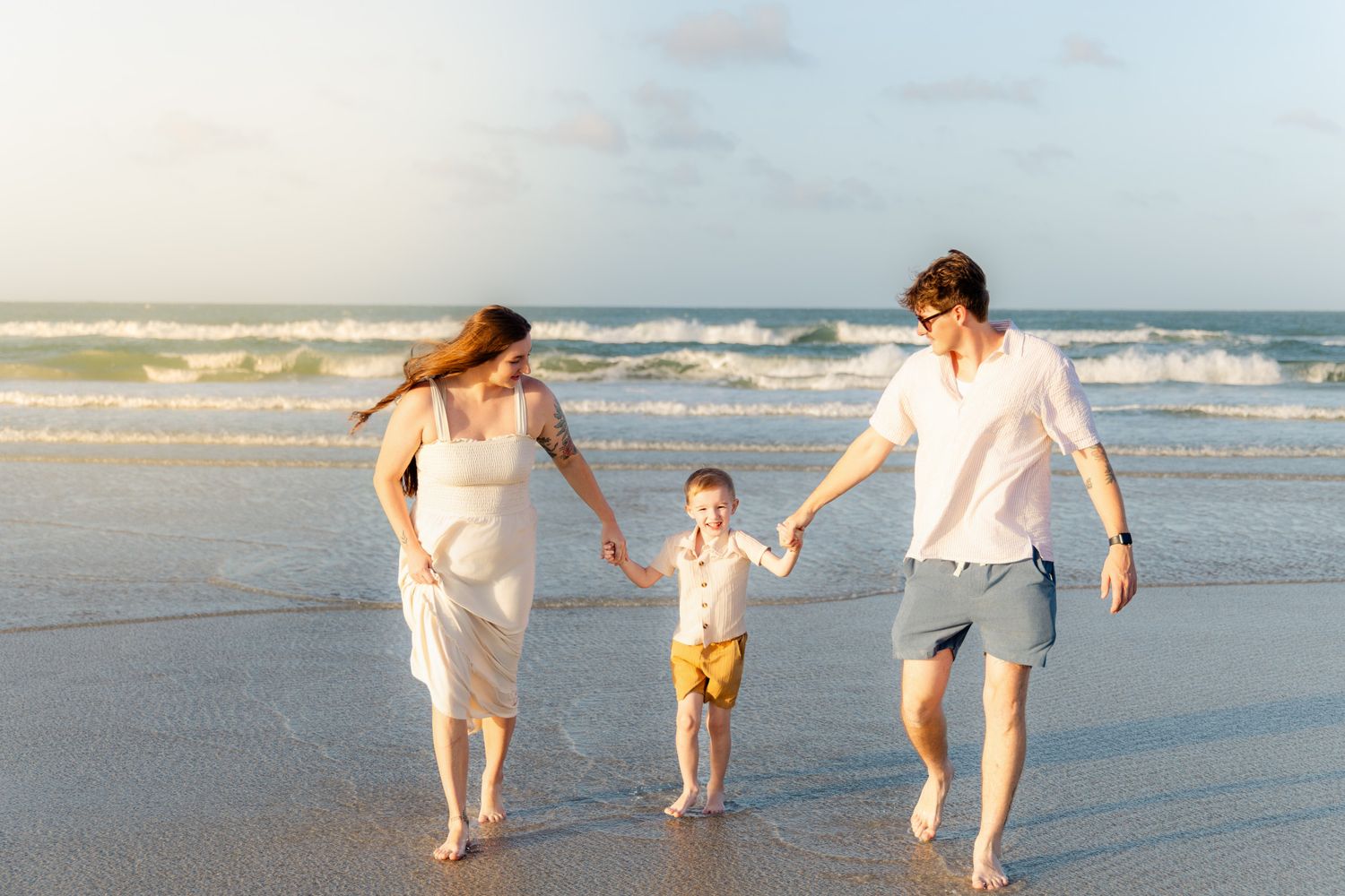 Family of three holding hands walking on a sunny beach near ocean waves.